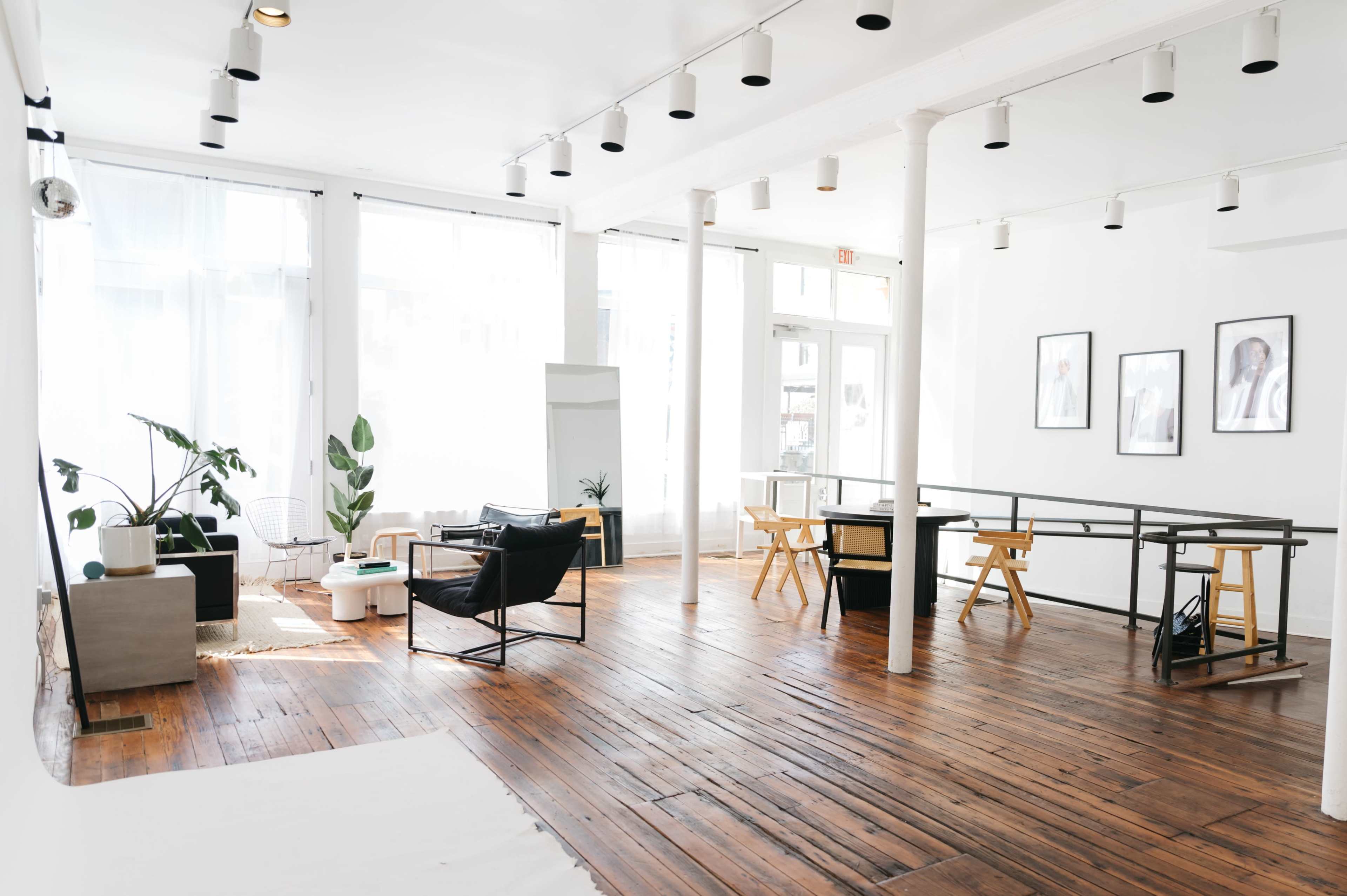 The image features a spacious, well-lit interior with wooden floors, minimalist furniture, and large windows.