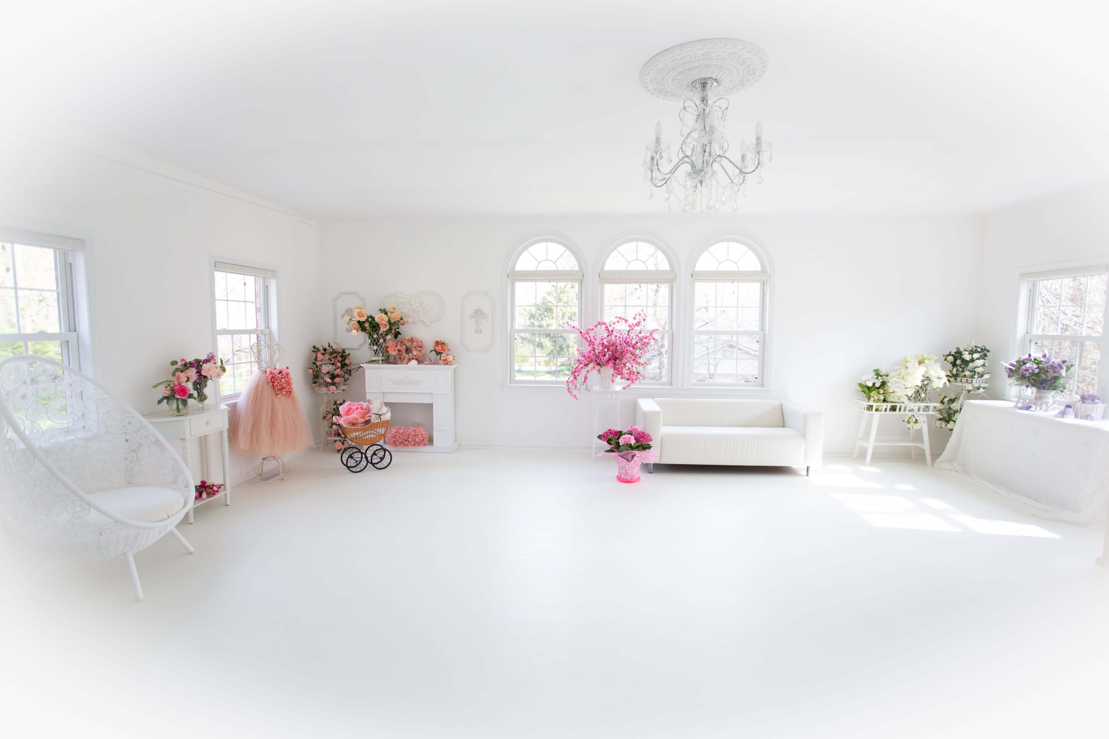 The image depicts a bright, airy room with white walls and elegant decor, featuring large windows, a chandelier, and various floral arrangements.