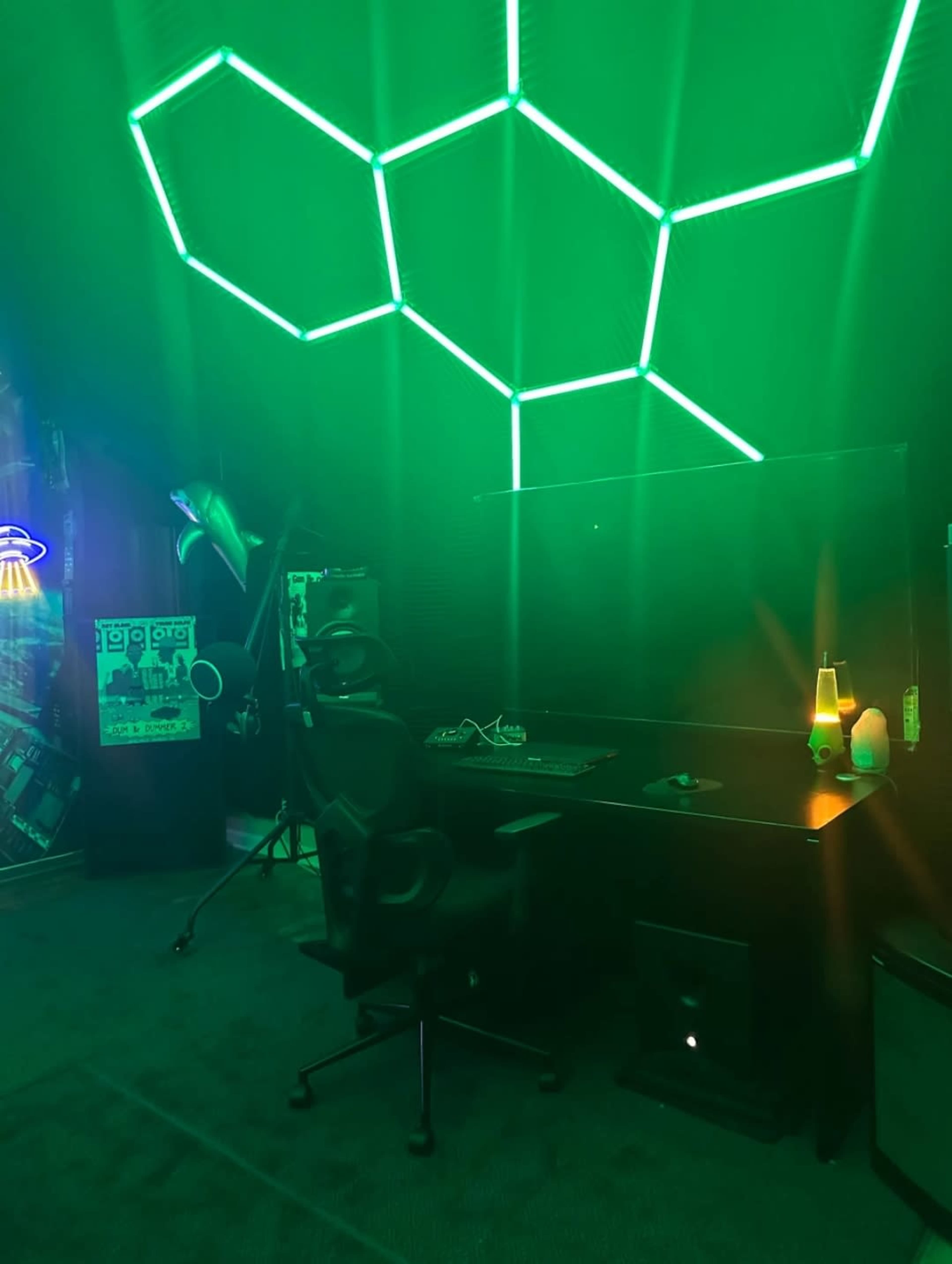 The image shows a dimly lit room with a green neon light pattern on the ceiling and a black desk with a chair, accompanied by various equipment and decor.