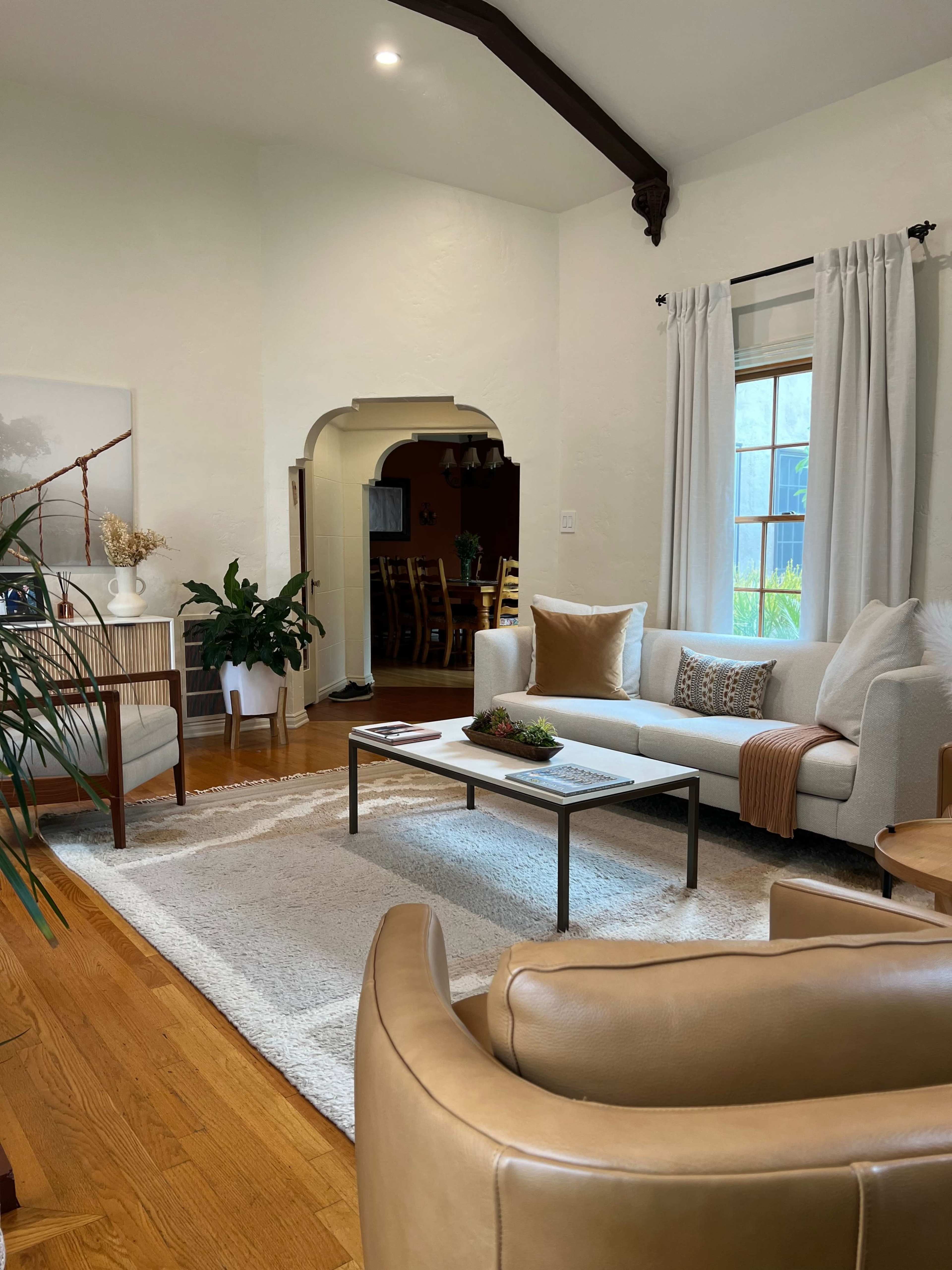 A cozy living room features a white sofa, wooden furniture, and green plants, with an archway leading to another room in the background.