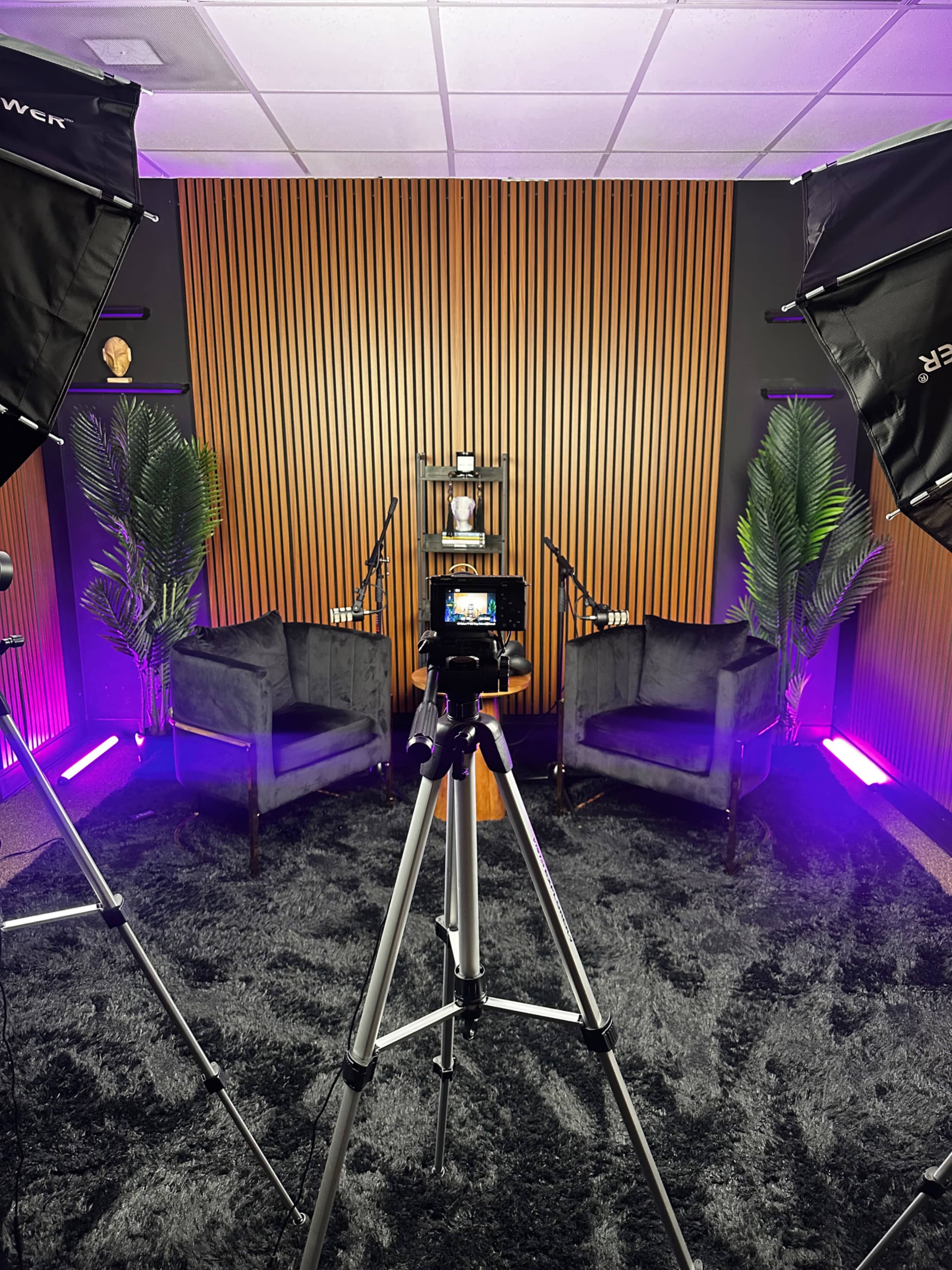 A modern podcast studio features two black chairs, a shelf with decor items, and vibrant lighting, surrounded by plants and camera equipment.