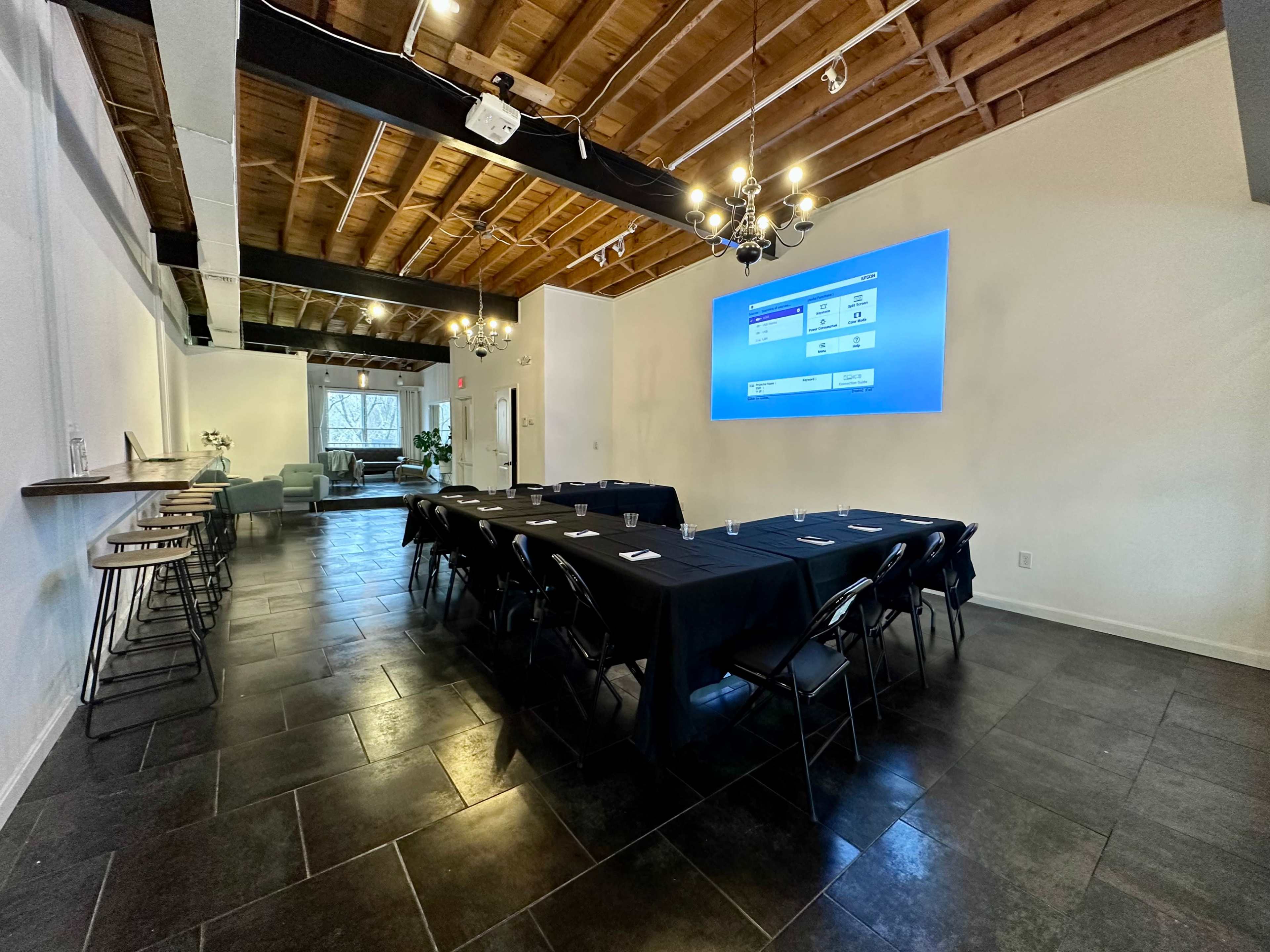 A conference room is set up with a long table covered in a black tablecloth, surrounded by chairs, and a projector screen displaying information on the wall.