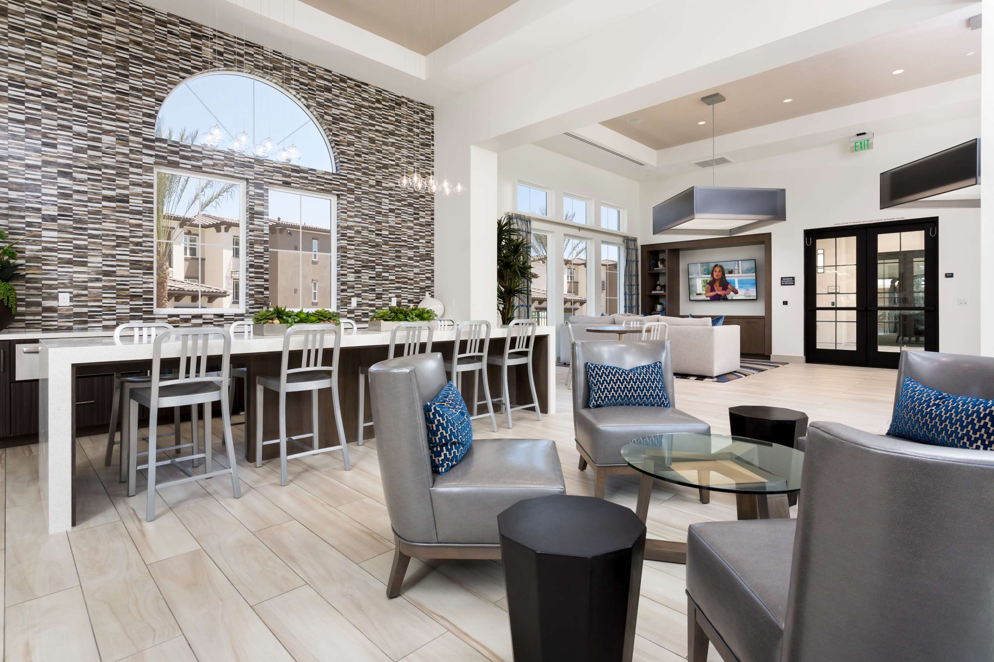 The image shows a modern common area featuring a bar with high stools, a lounge seating arrangement, and large windows allowing natural light to enter.