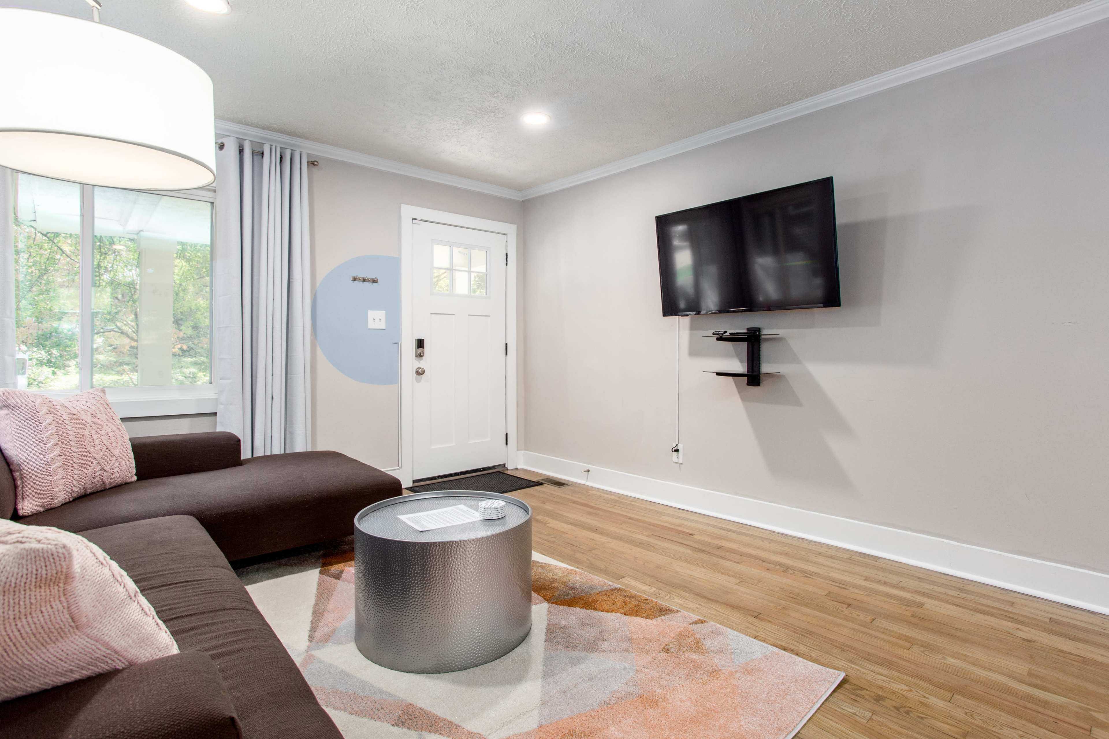 The living room features a dark brown sectional sofa, a round metallic coffee table, and a wall-mounted television, with a door leading outside and windows allowing natural light.