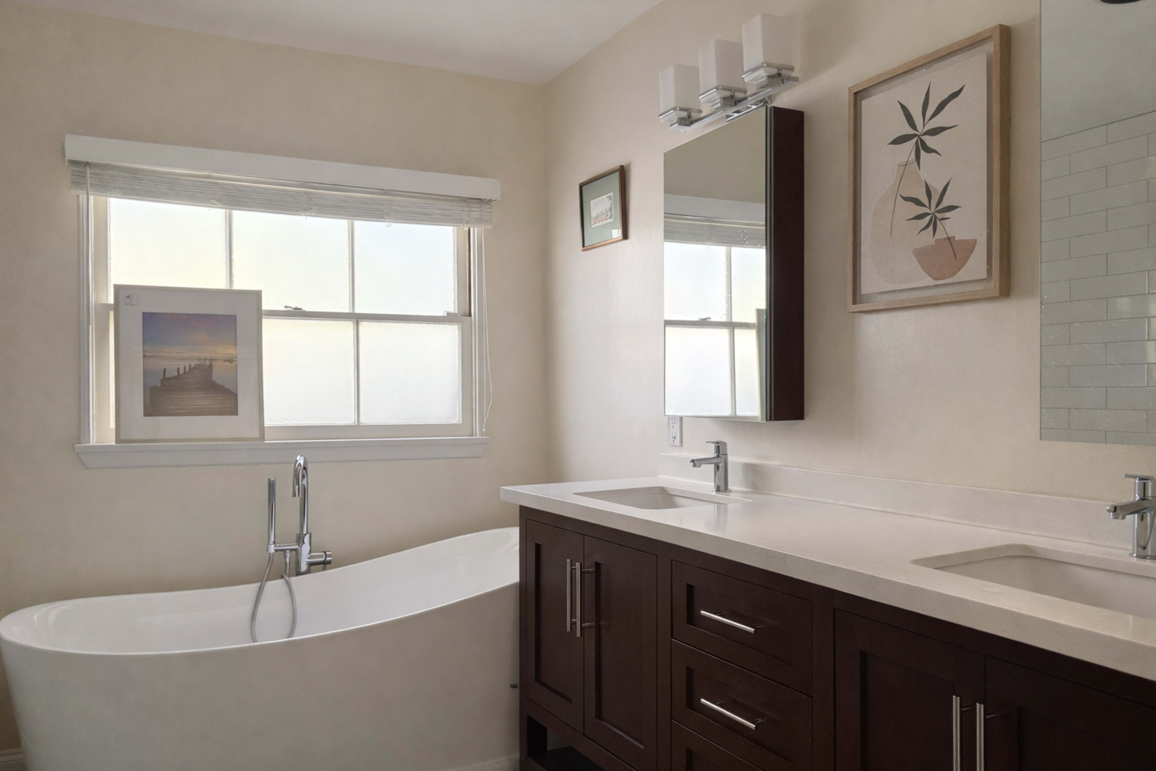 The image shows a modern bathroom featuring a freestanding bathtub, a double sink vanity with dark wood cabinetry, and framed artwork on the walls beside a large window.