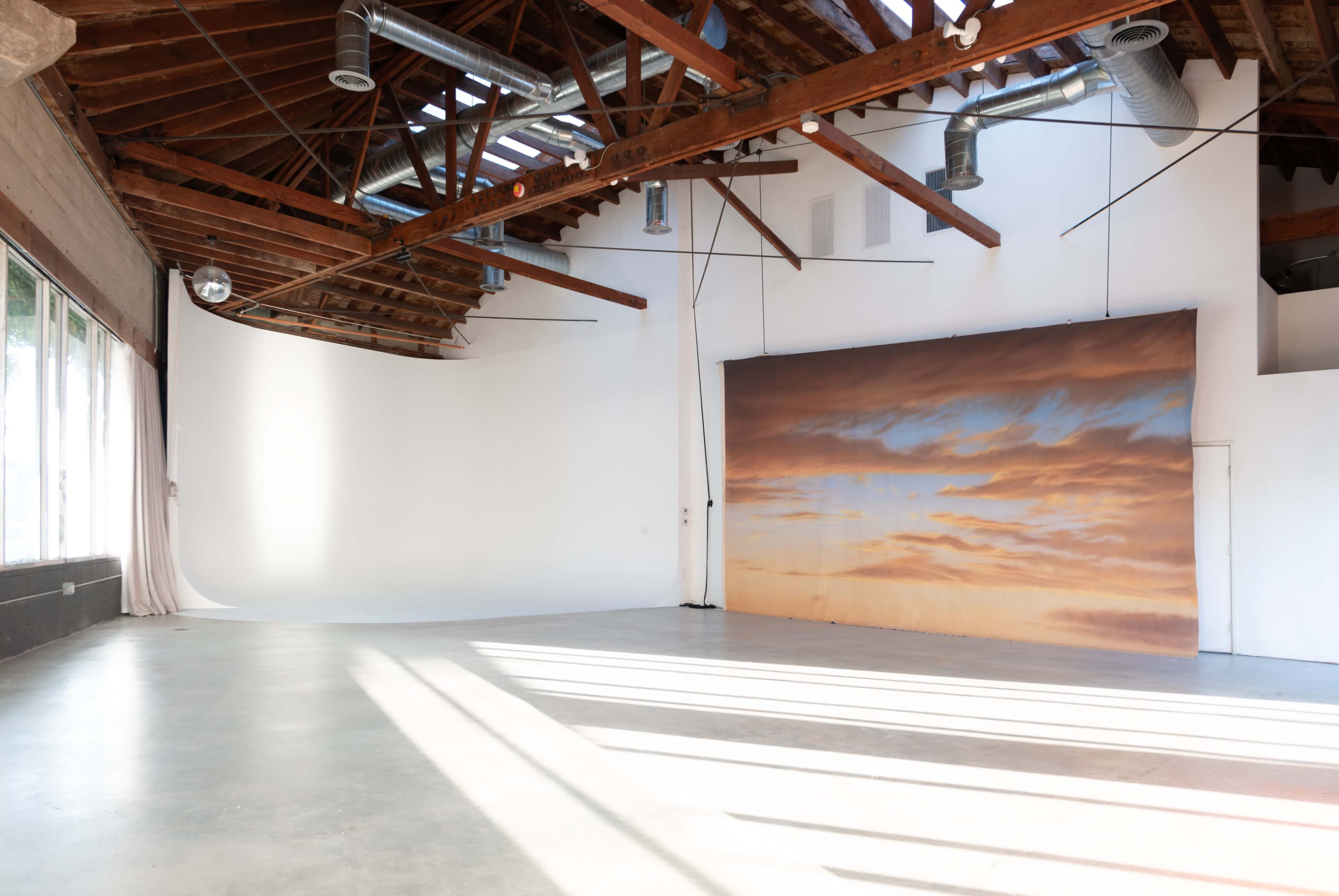 The image shows a spacious studio with exposed wooden beams, large windows letting in natural light, and a backdrop featuring a serene sunset scene.
