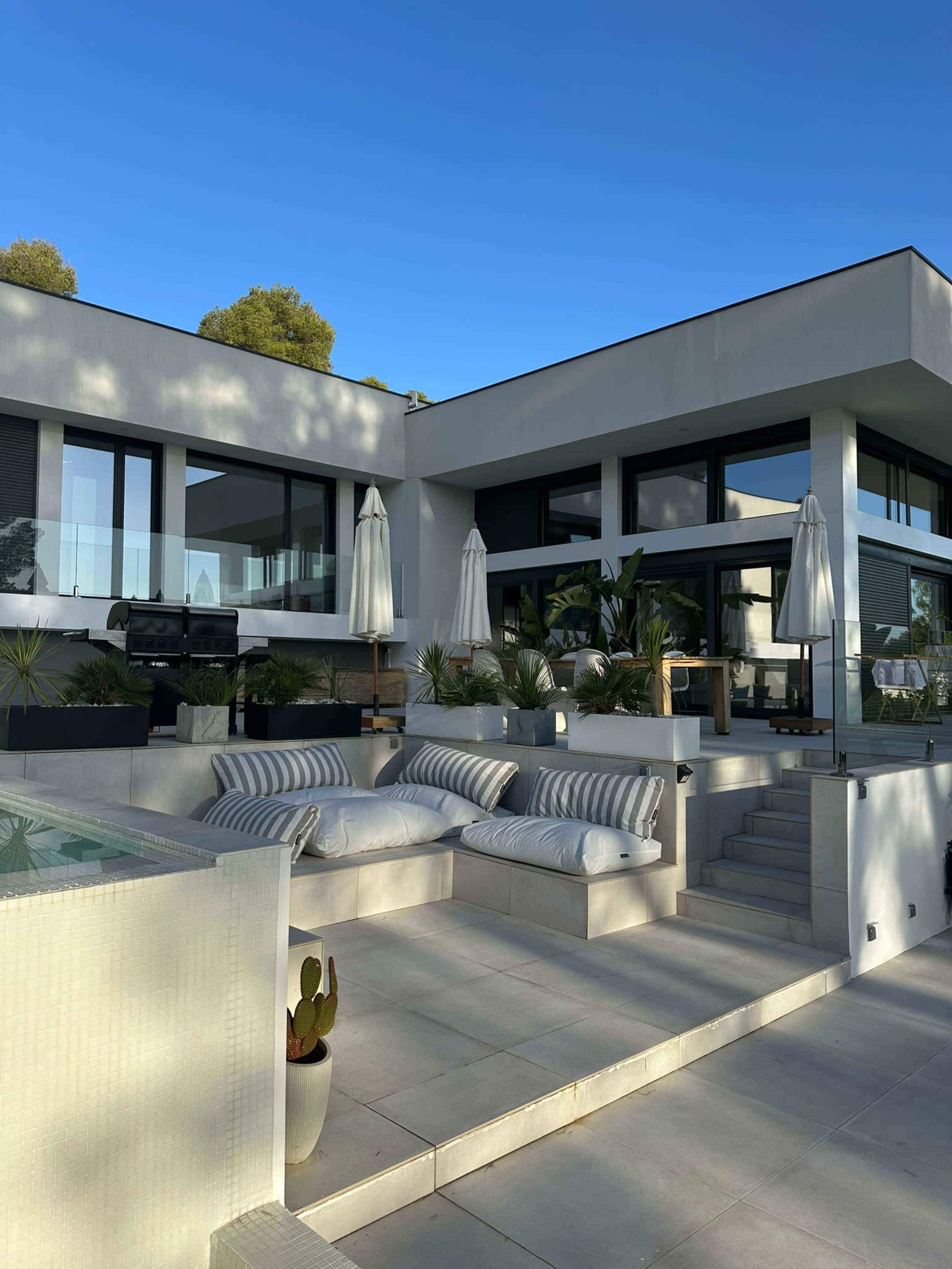 The image shows a modern, two-story house with large glass windows, an outdoor seating area, and lush greenery surrounding the patio.
