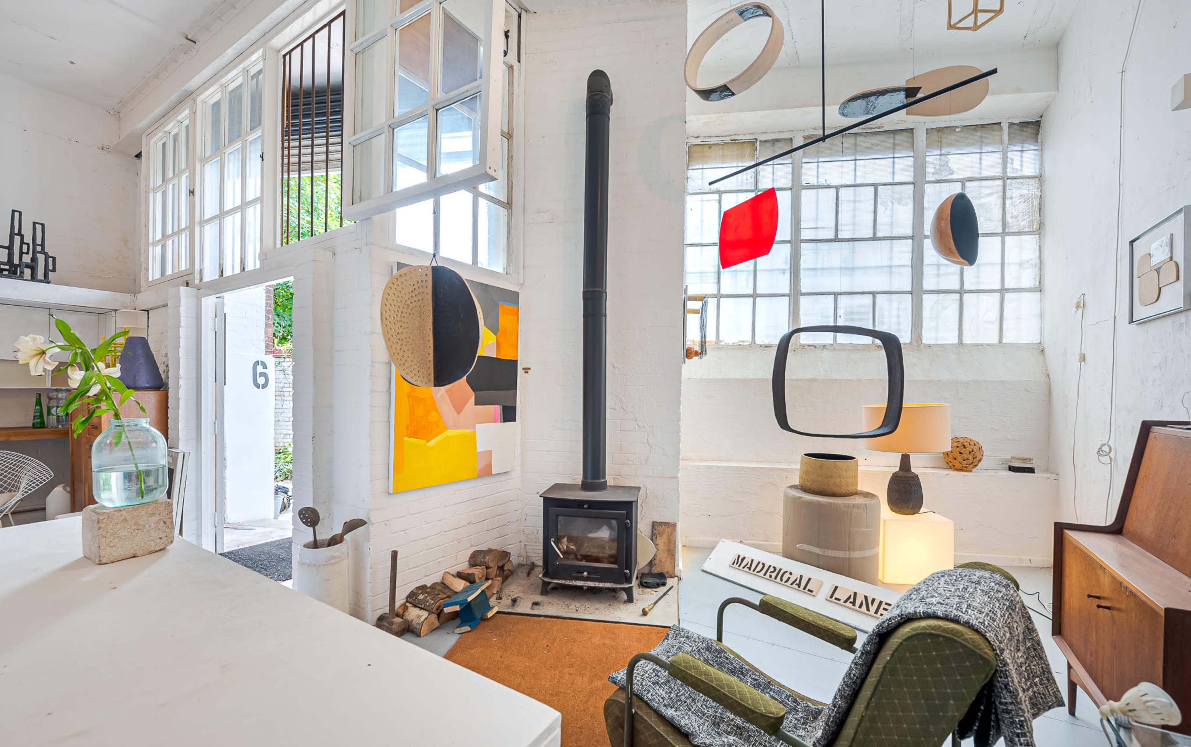 The interior of a modern studio features a wood stove, colorful wall art, and various hanging light fixtures, with large windows allowing natural light to enter.