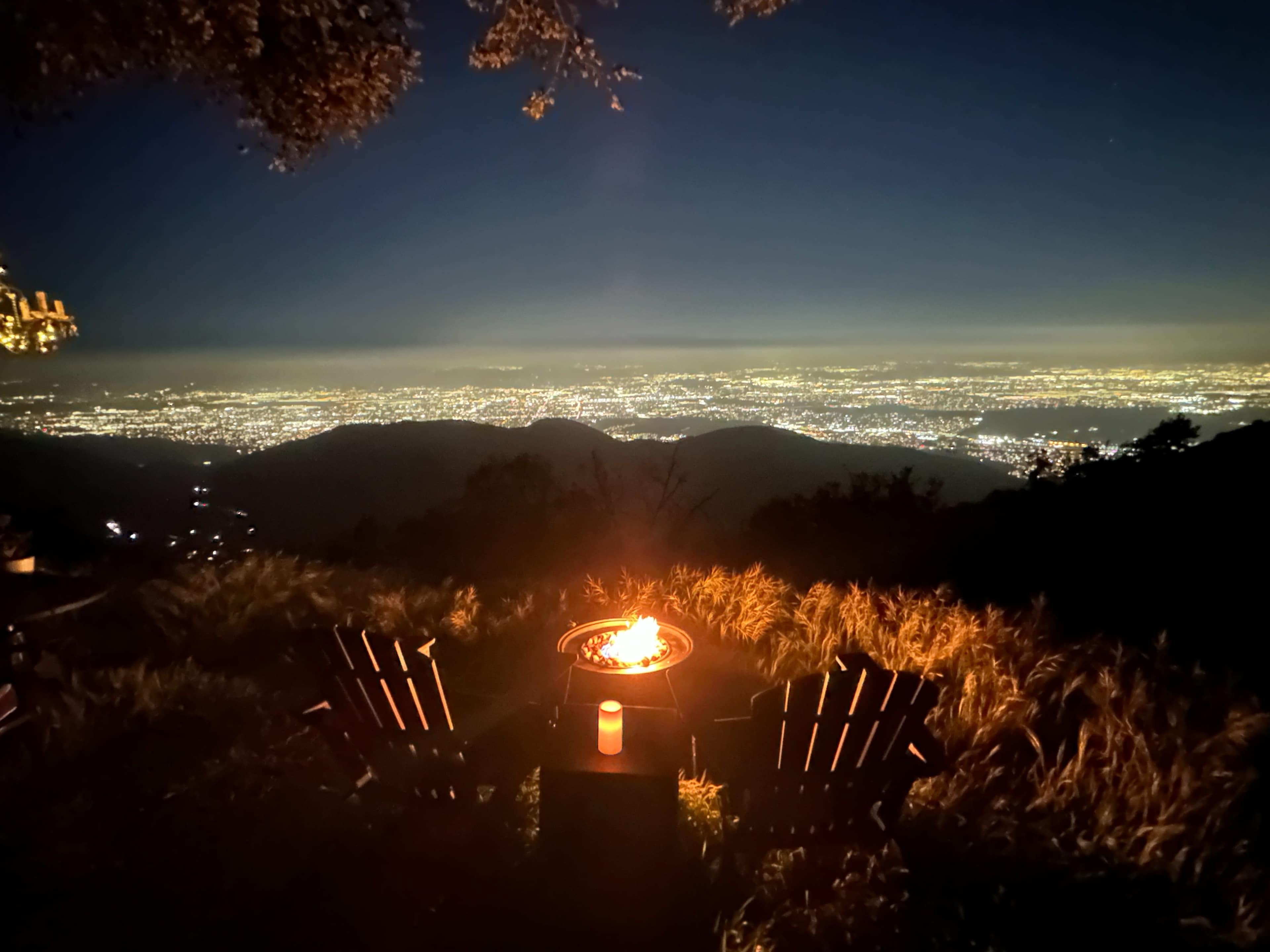 A fire pit surrounded by two chairs overlooks a lit cityscape at night.
