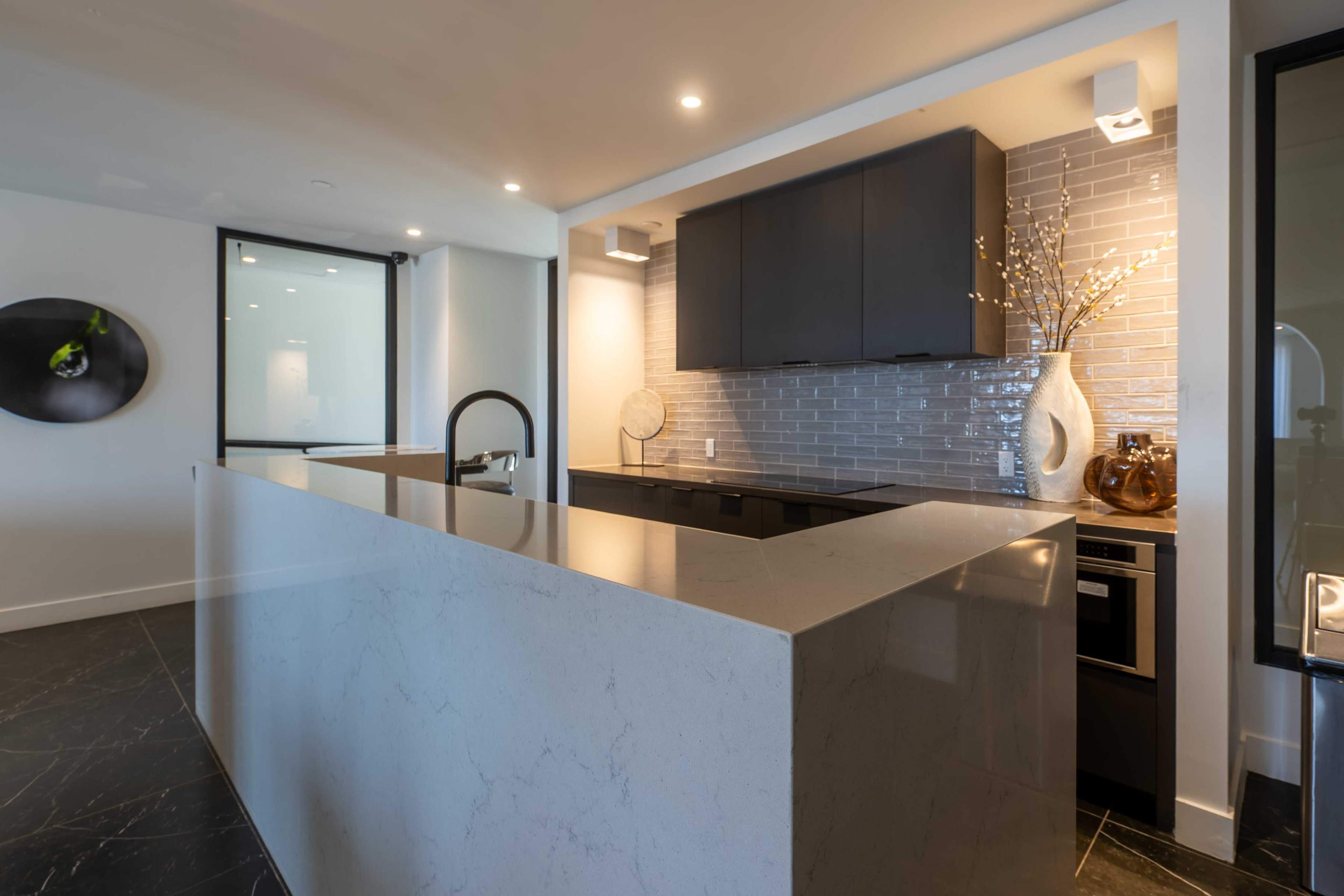 The image shows a modern kitchen featuring a large island with a marble countertop, sleek dark cabinetry, and a stylish backsplash.