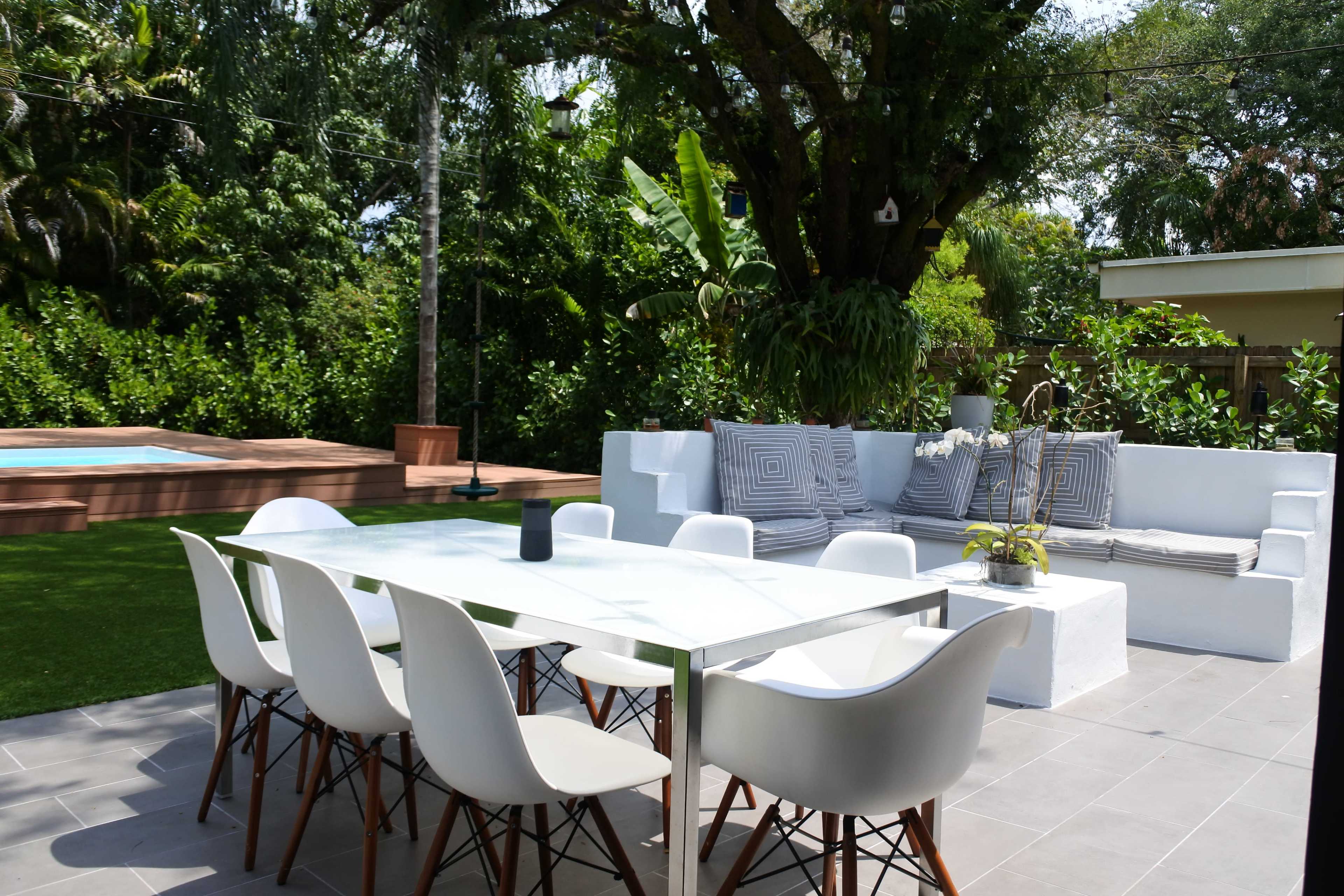 A modern outdoor dining area features a glass table surrounded by white chairs, with a plush seating arrangement nearby and a swimming pool in the background.