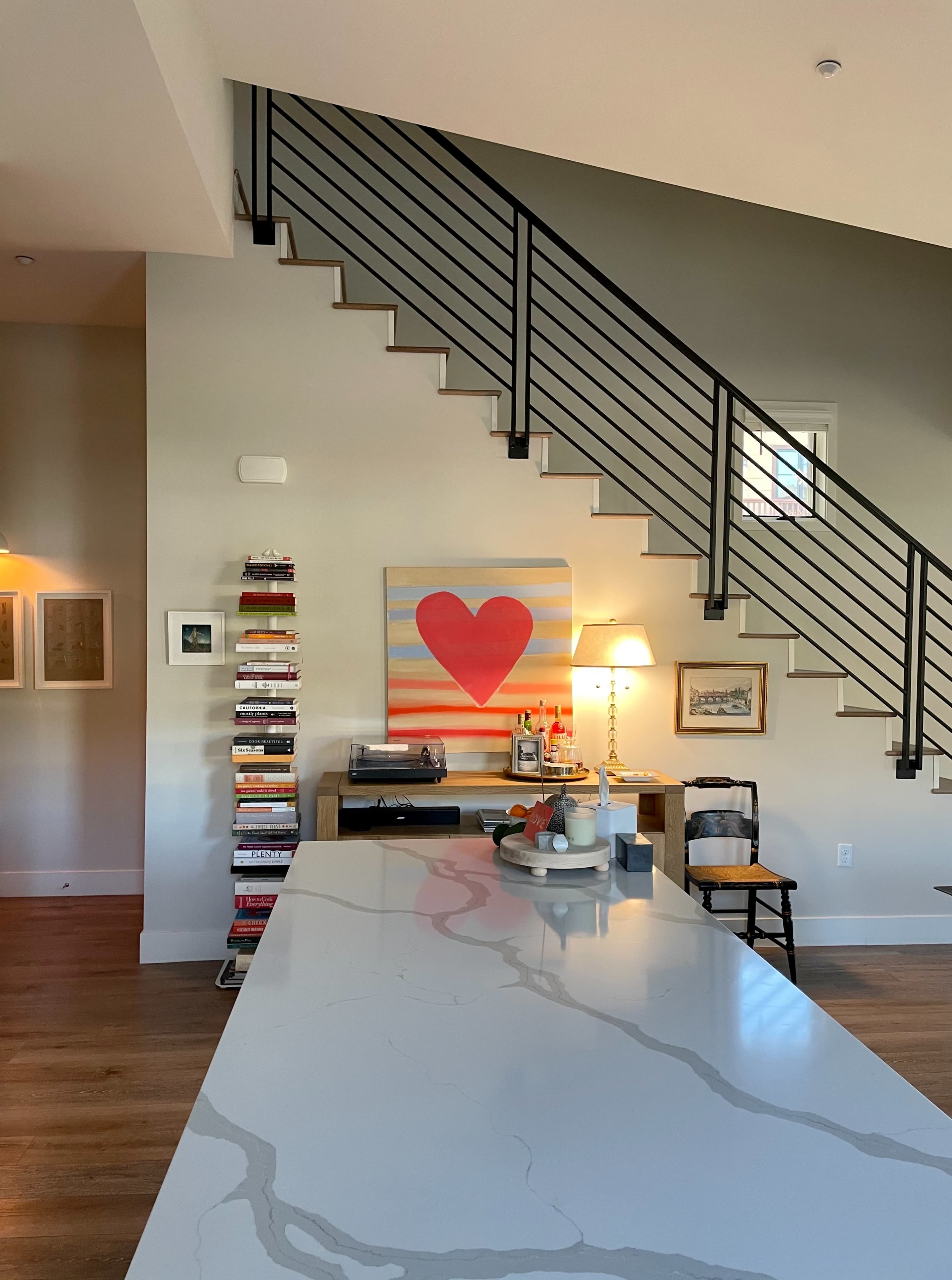 A modern interior features a staircase with black railings, a large heart painting on the wall, and a sleek kitchen island with books and decor items.