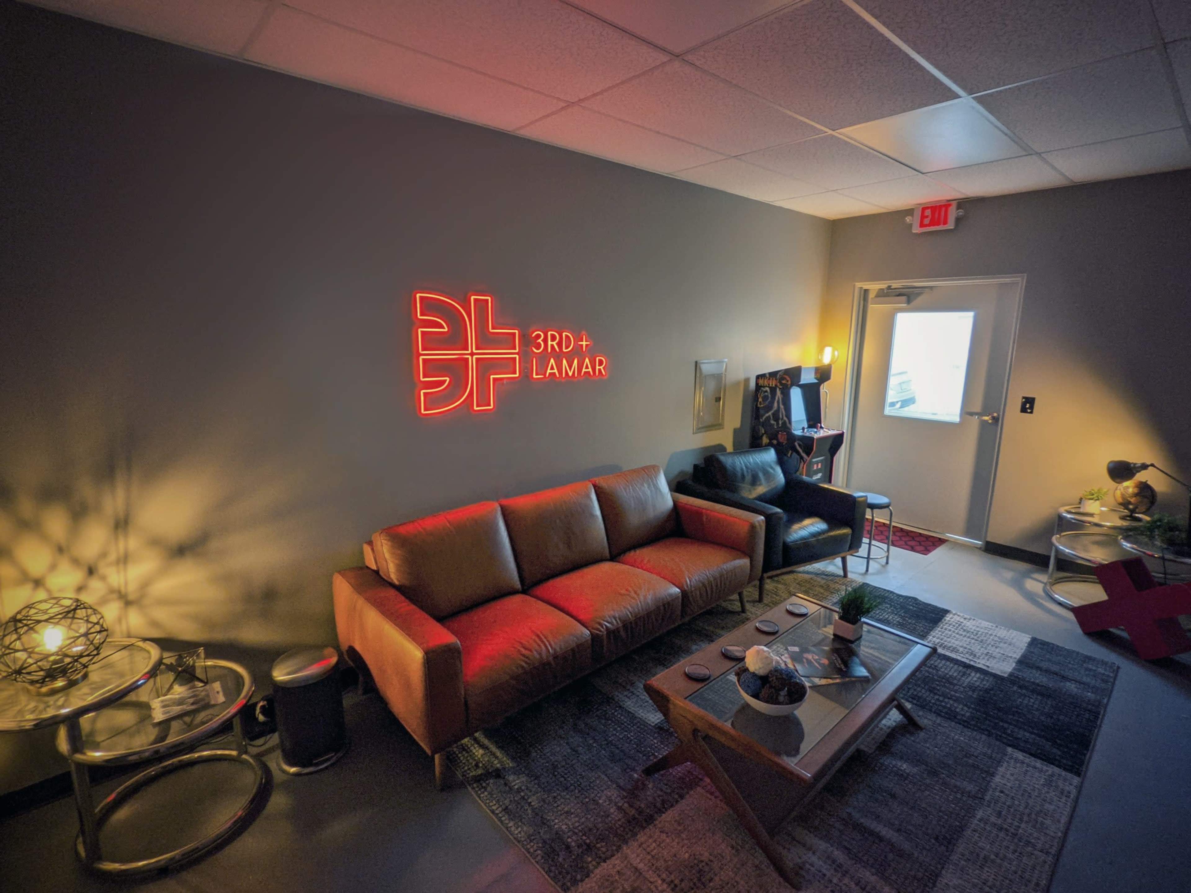 The image shows a modern lounge area with a glowing neon sign that reads "3RD + LAMAR," featuring a brown sofa, a black chair, and various decorative items on a table.