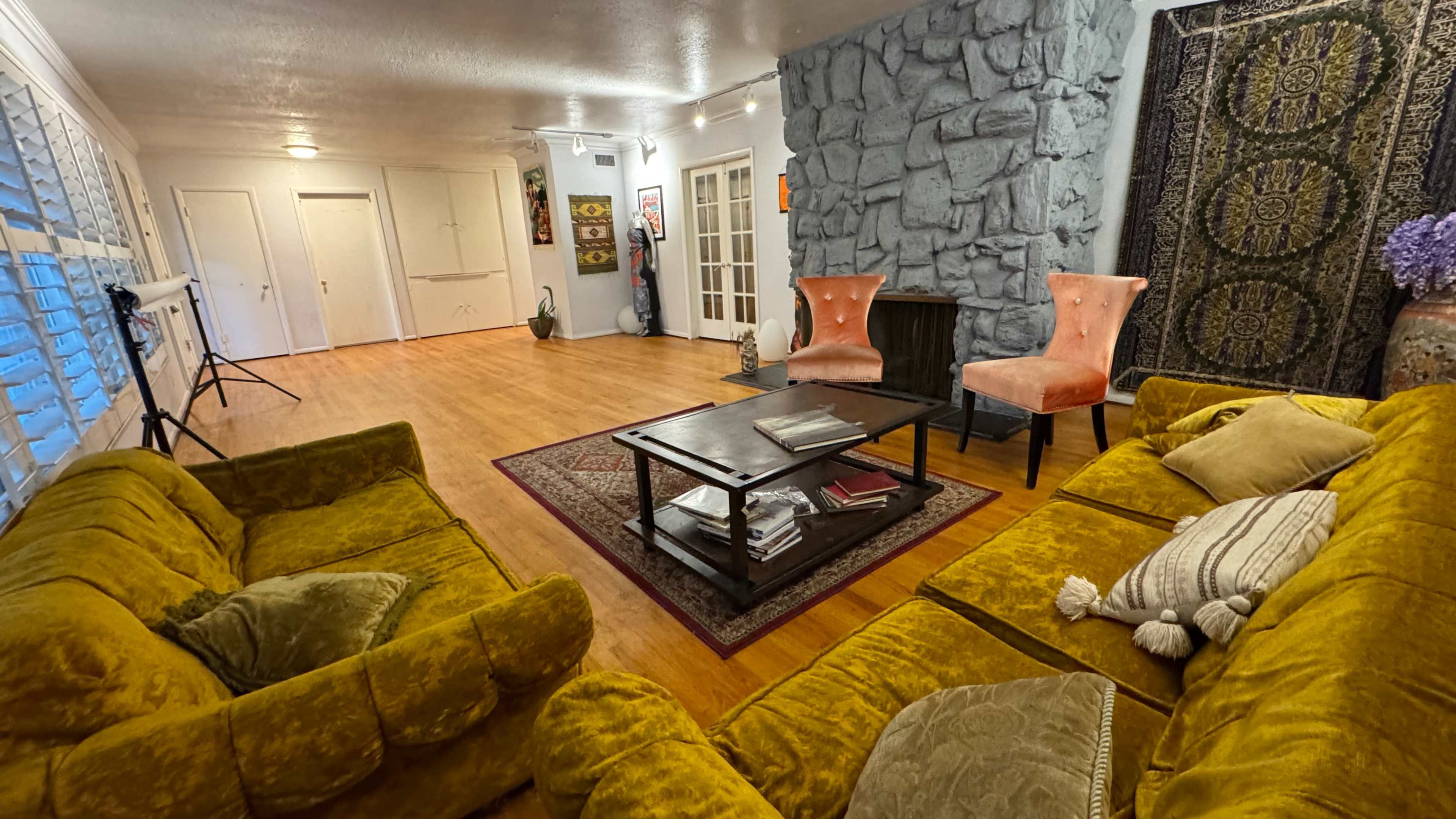 The image shows a cozy living room with a stone accent wall, two pink chairs, a wooden coffee table, and a large yellow sectional sofa.