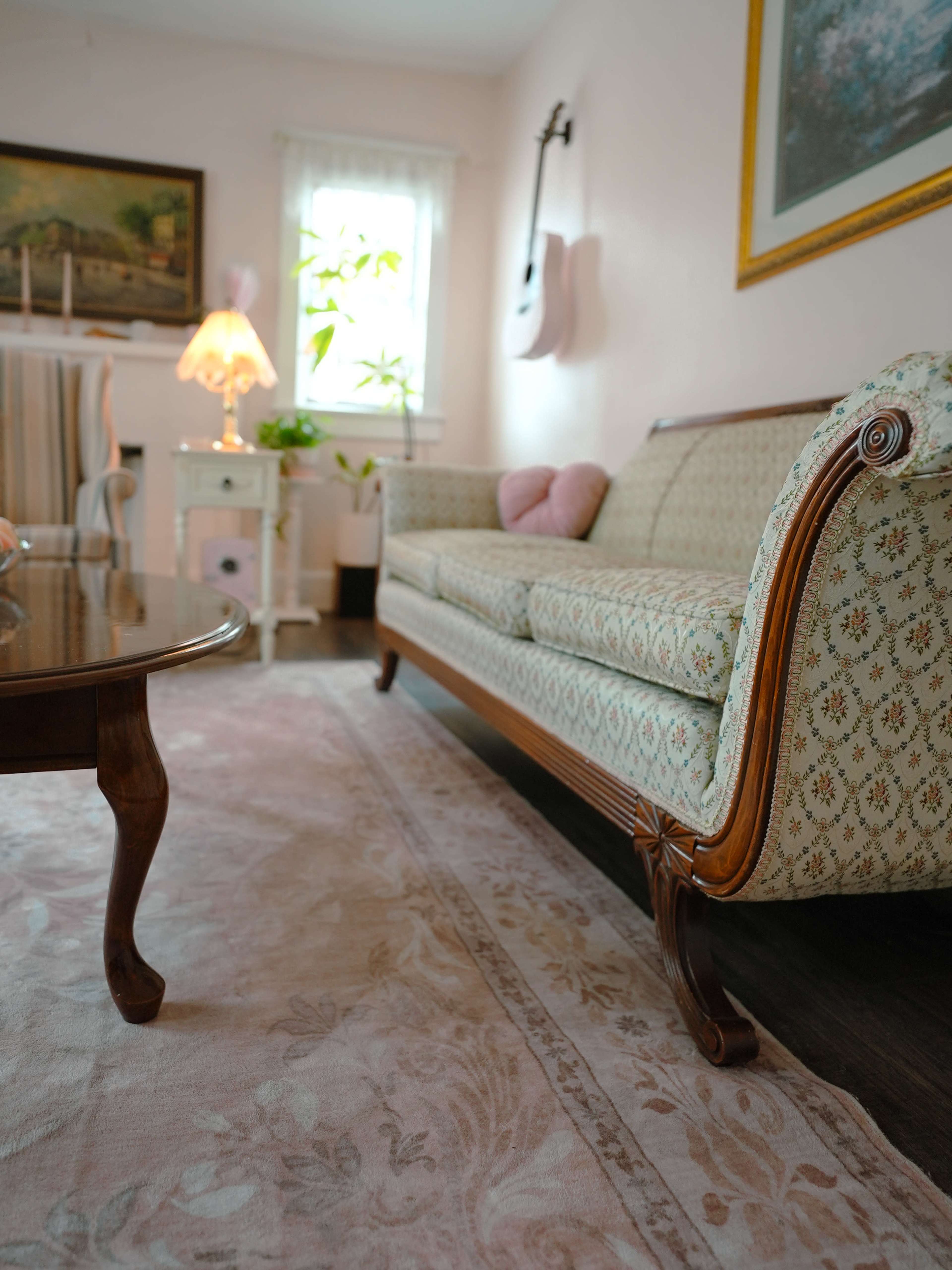 The image shows a living room with a patterned sofa, a wooden coffee table, a lamp, and decorative elements including a guitar on the wall and a plant by the window.