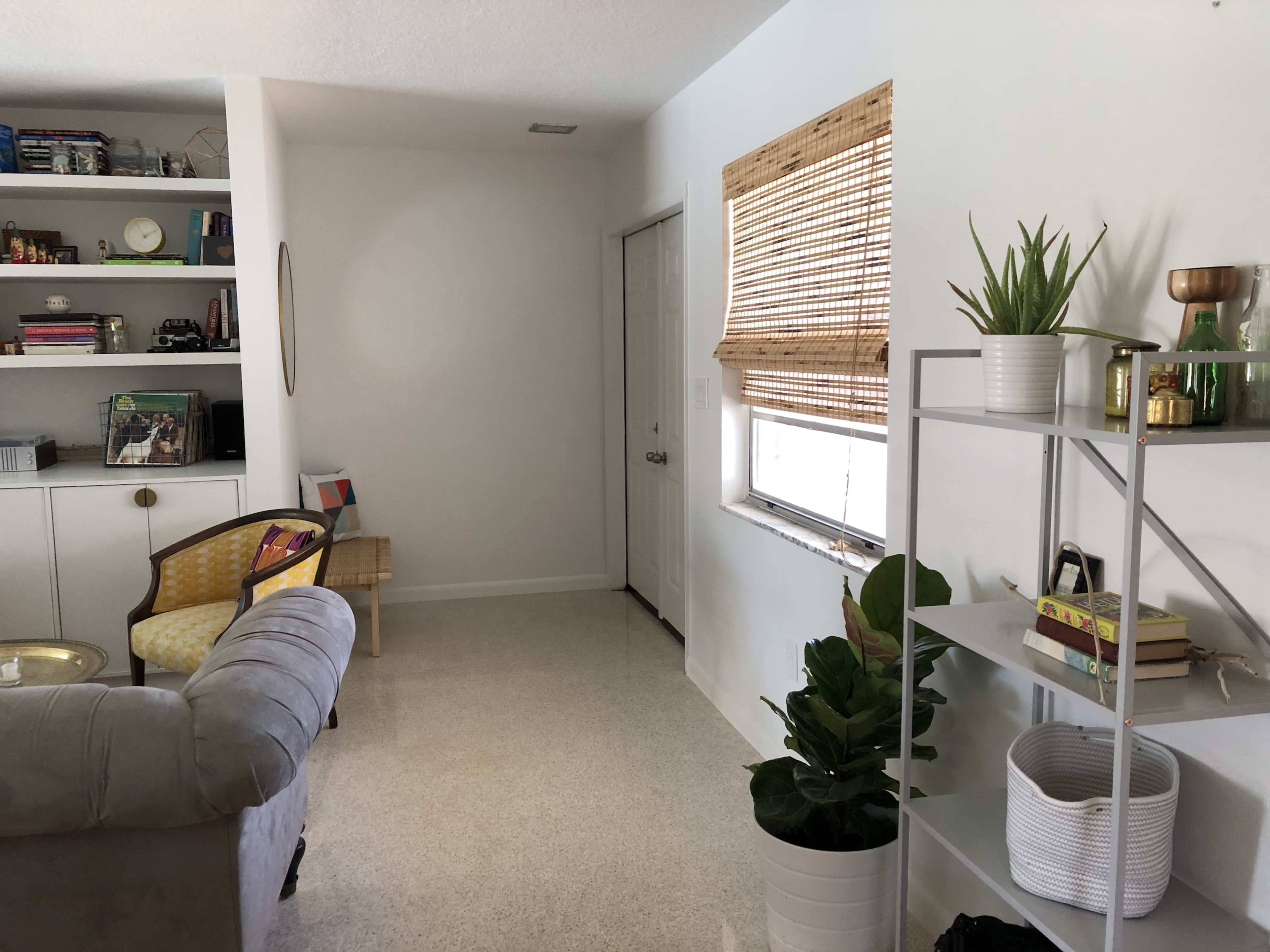 The image shows a bright, minimalistic living space with a gray sofa, a bookshelf with decorative items, a door leading to another room, and plants placed around the area.
