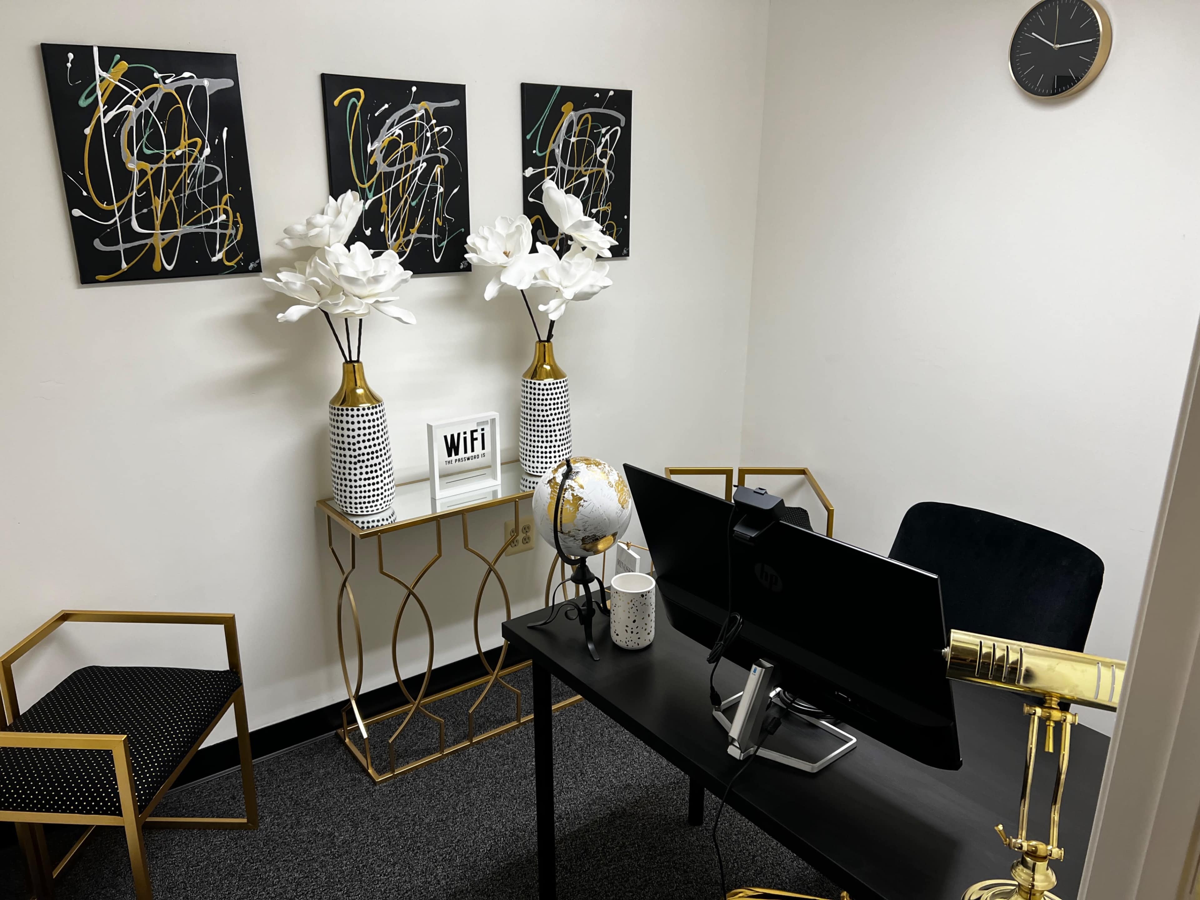 A small office space featuring a black desk with a computer, decorative chairs, and wall art, along with floral arrangements and a globe on display.