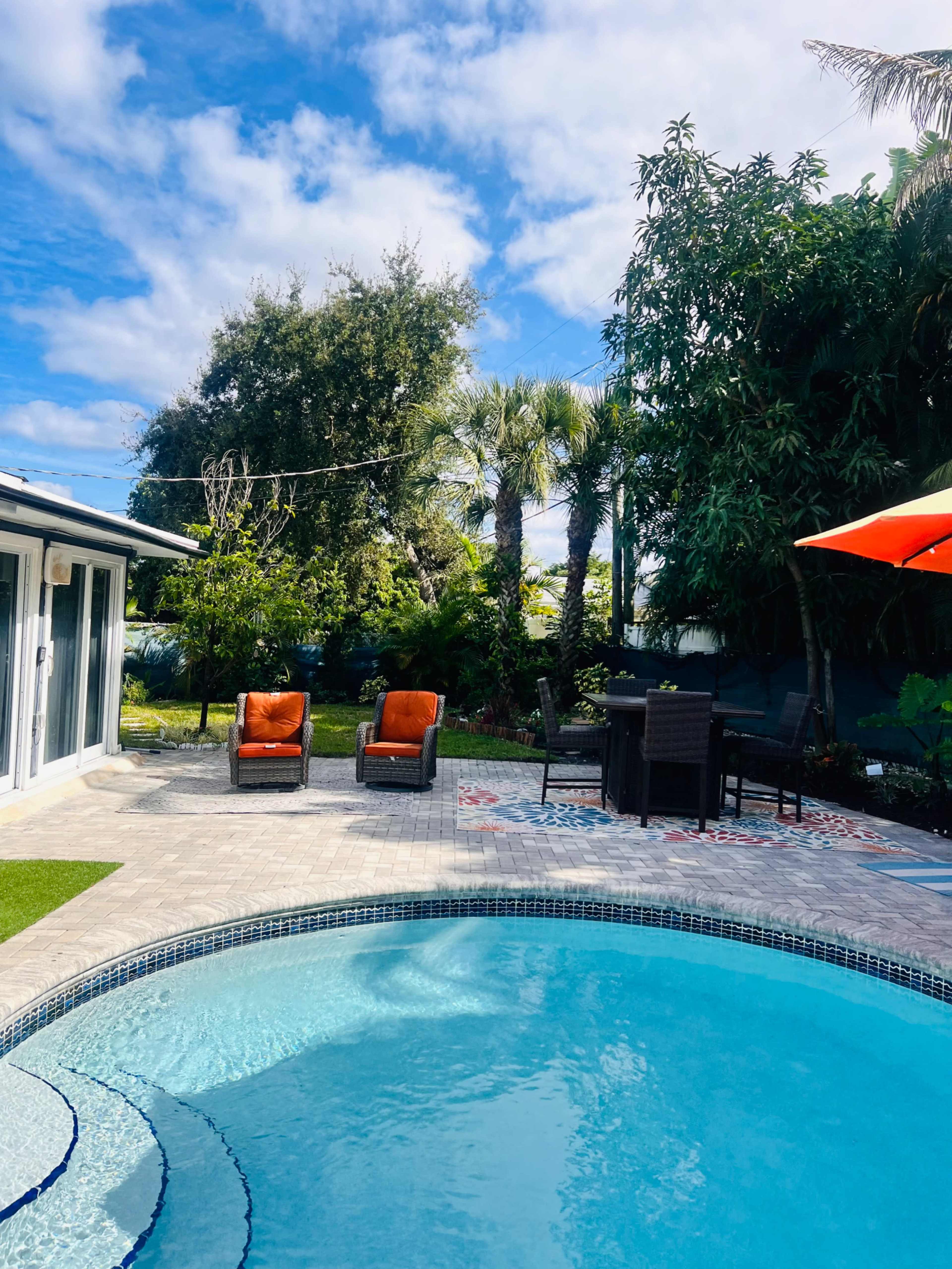 The image depicts a bright outdoor pool area with two orange chairs, a dining table, and lush greenery in the background.
