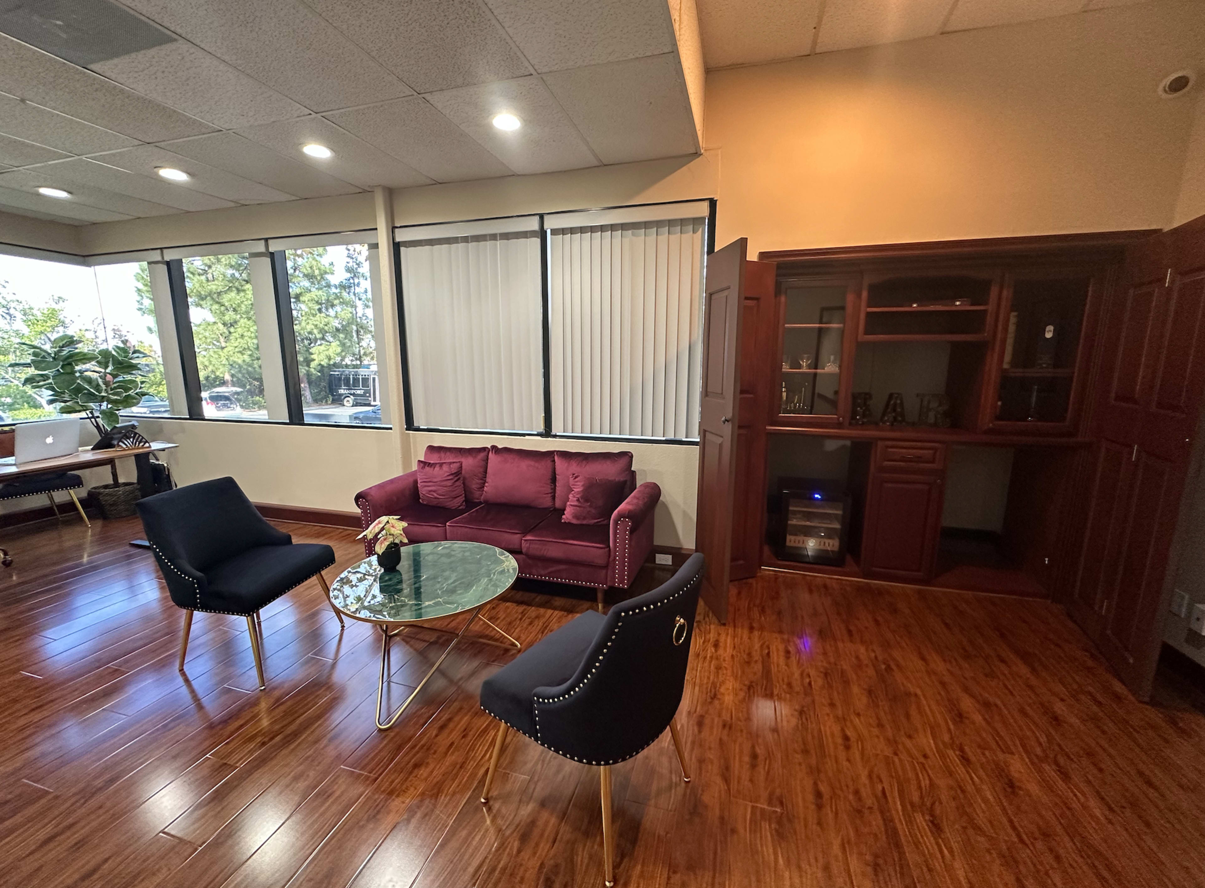 A modern office seating area with a burgundy couch, two black chairs, a round glass table, and a wooden cabinet against the wall.