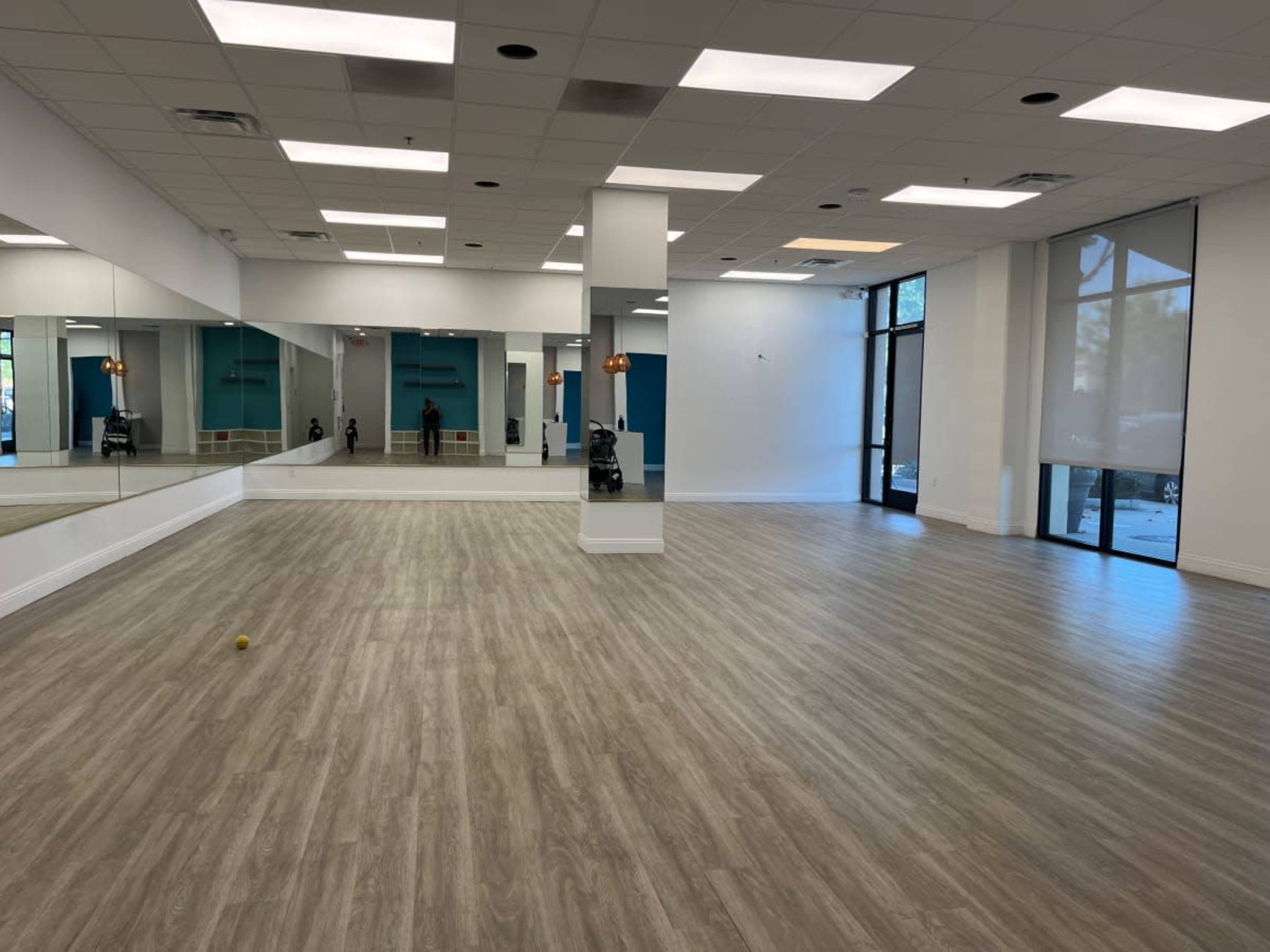 A spacious, empty room with wooden flooring, large mirrors on one wall, and floor-to-ceiling windows allowing natural light to enter.