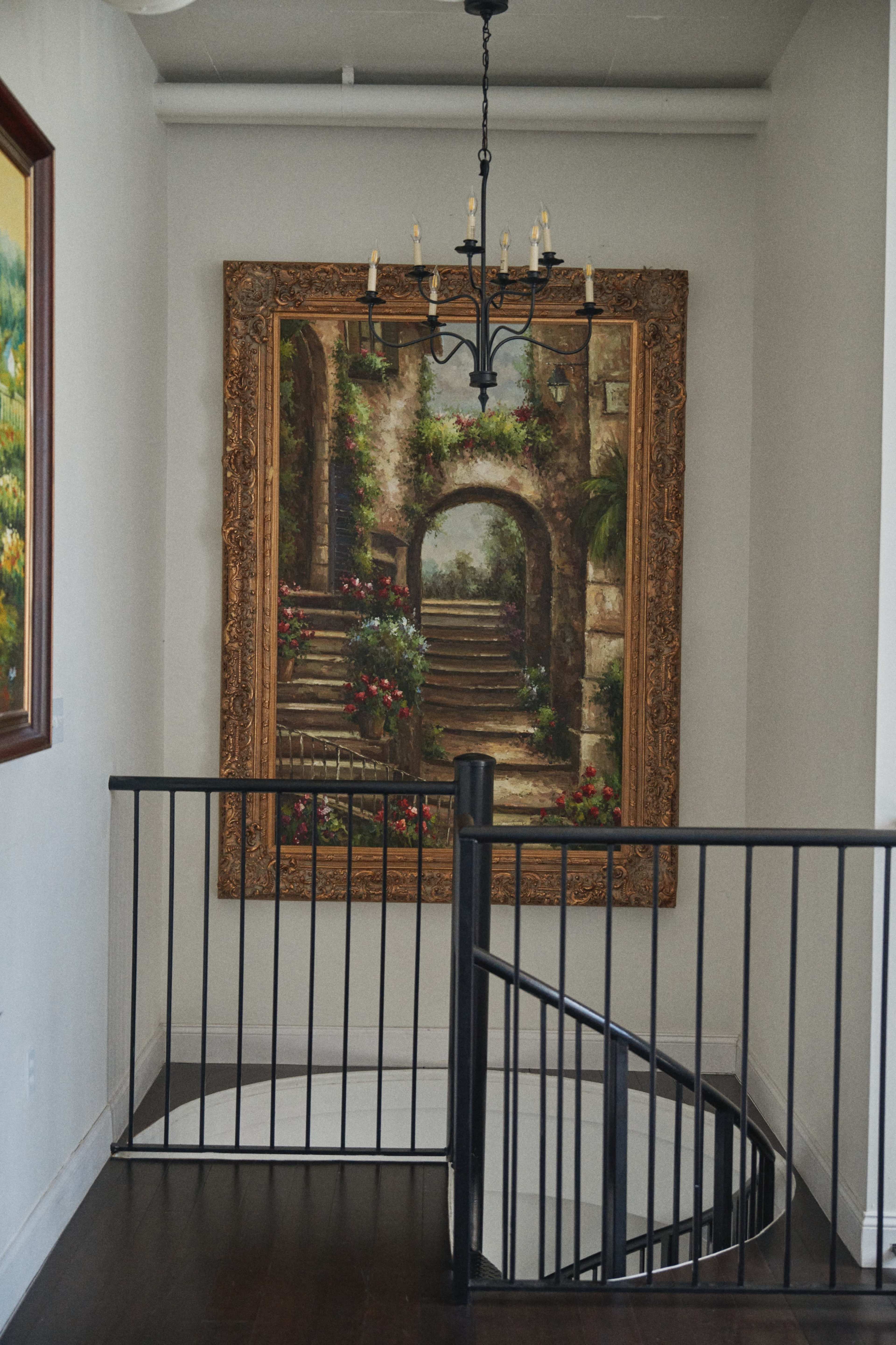 A large framed painting of a stairway adorned with flowers hangs on a wall near a spiral staircase and a chandelier.