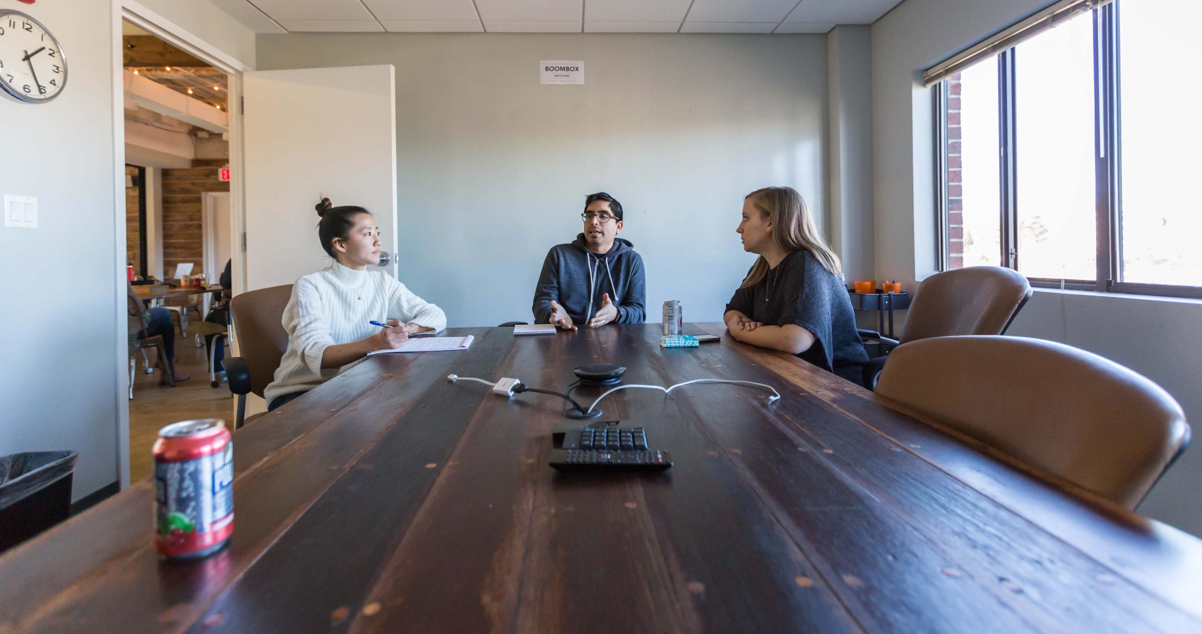 Three people are seated around a wooden conference table in an office meeting room, engaged in discussion.