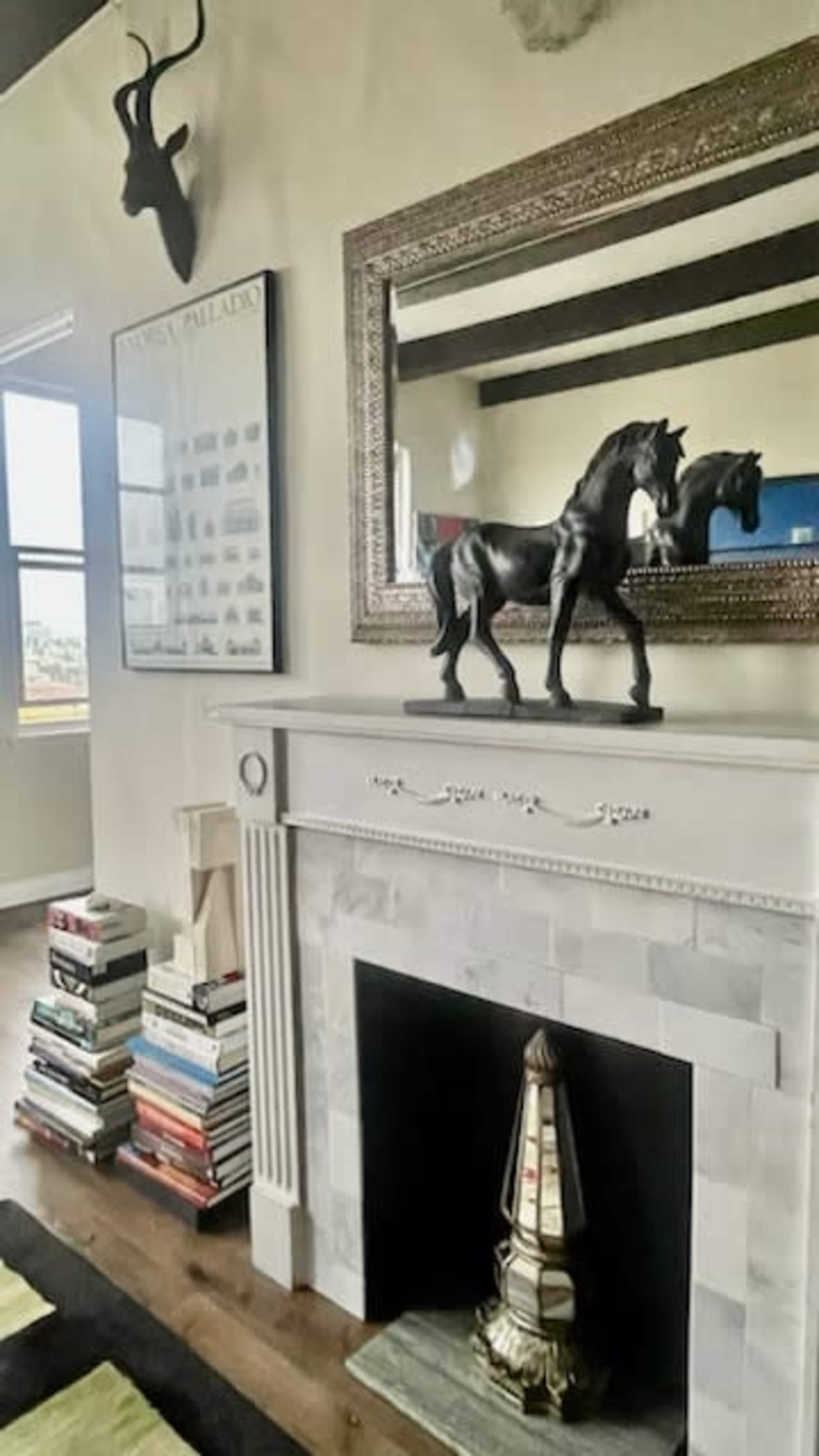 A modern living room features a fireplace with a decorative horse sculpture atop the mantel, surrounded by a tall stack of books and a mirrored wall with a framed print.