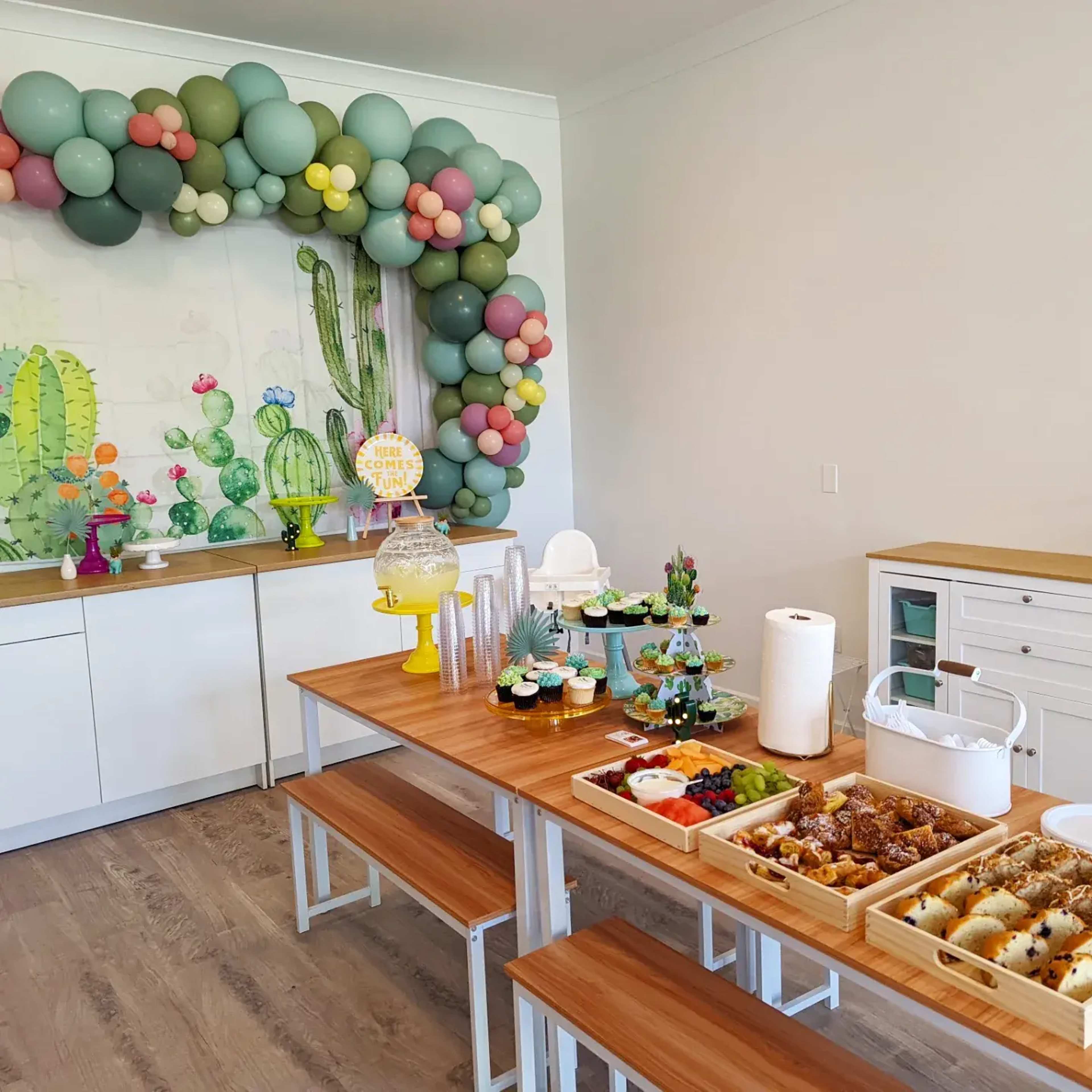 A decorated party space with a balloon arch, a table set with a variety of food items, and wall art featuring cacti.