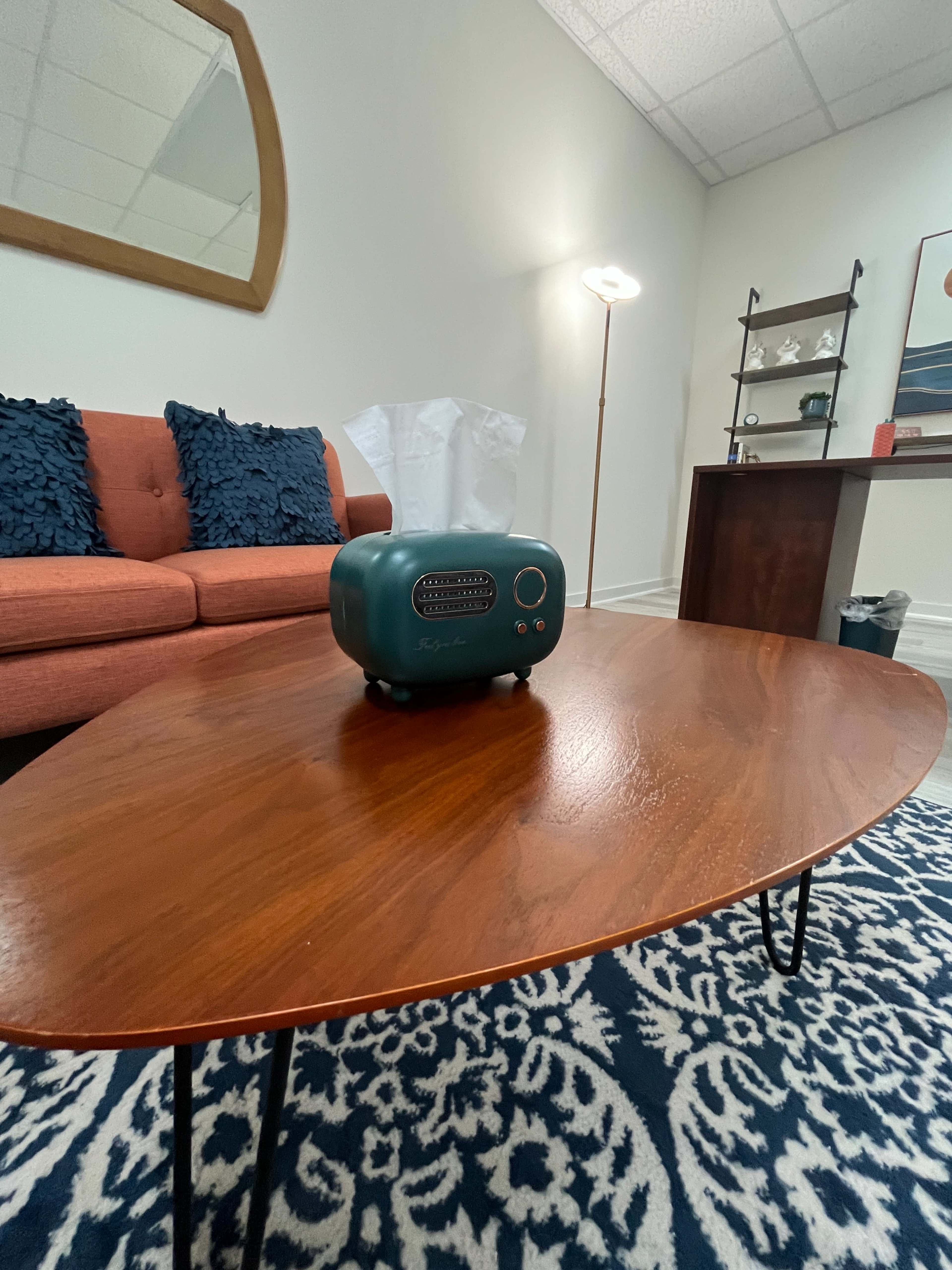 A vintage teal radio sits on a wooden coffee table in a modern living room with a coral sofa and decorative items on a shelf.