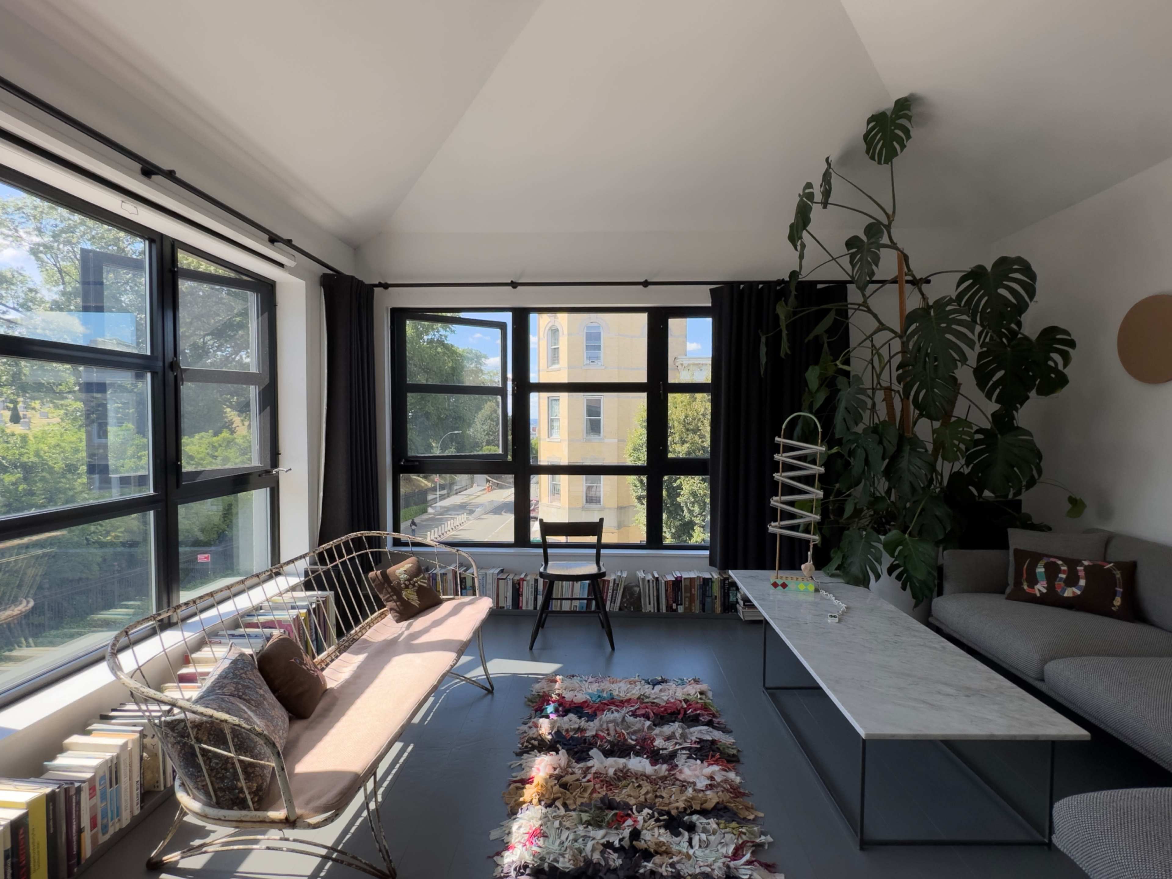 A sunlit living room features large windows, a minimalist gray sofa, a marble coffee table, a chair, and a pink bench adorned with cushions.