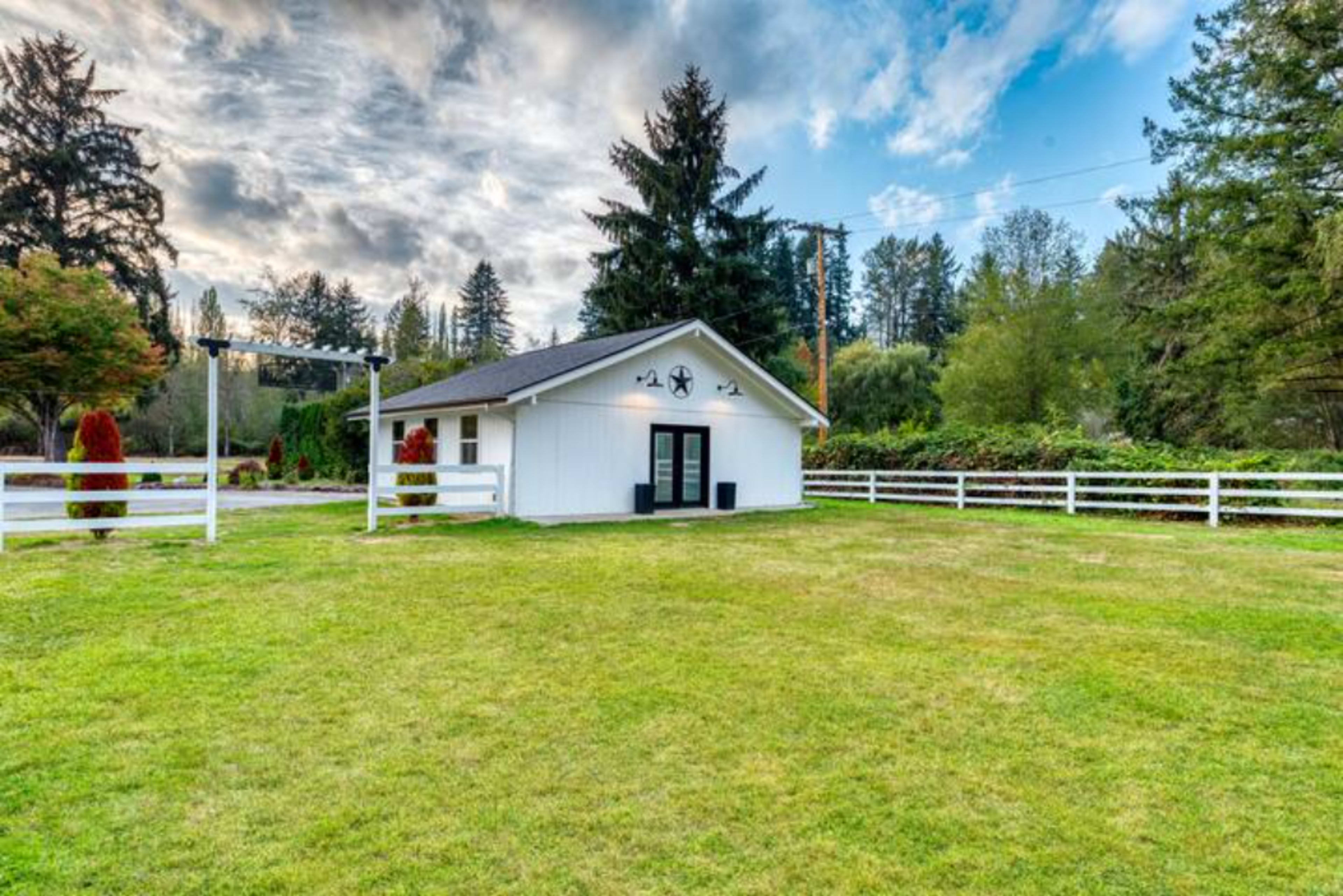 A small white building with a clock on the facade is surrounded by a green lawn and white picket fencing, under a partly cloudy sky.