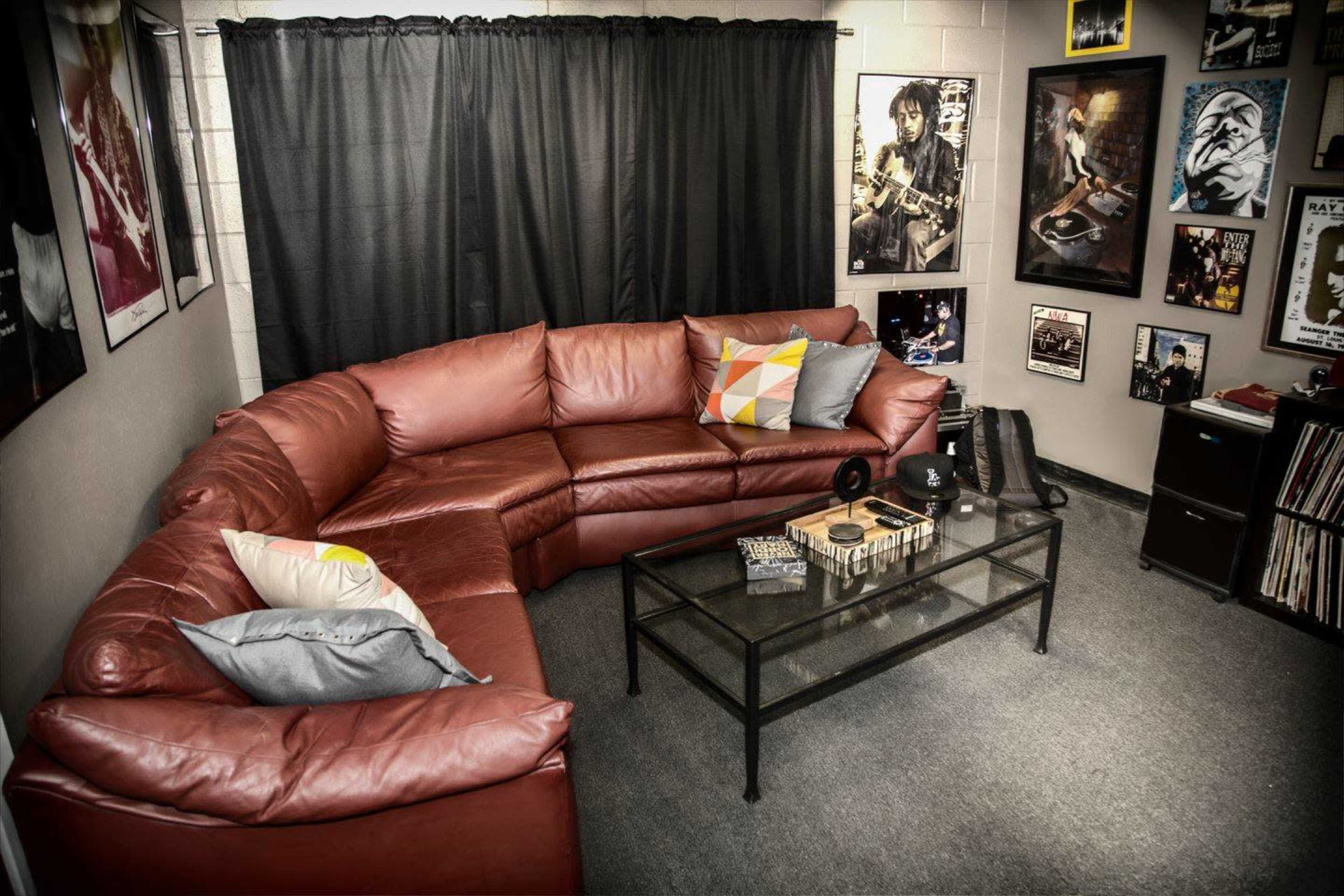 A red leather sectional sofa is arranged around a glass coffee table in a room featuring black curtains and walls adorned with various framed music posters.