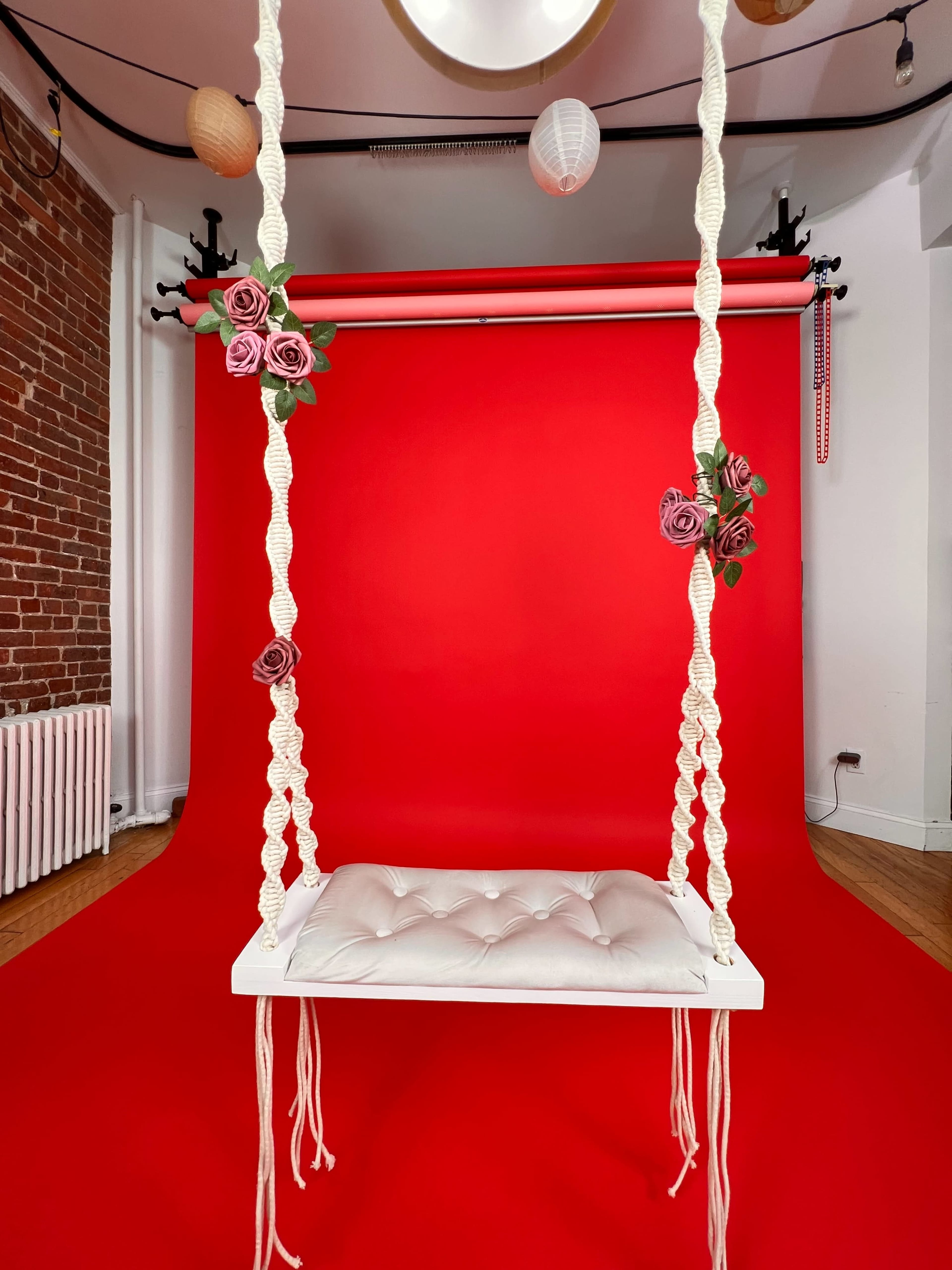 A white swing with a tufted seat is suspended from a red backdrop adorned with decorative flowers.
