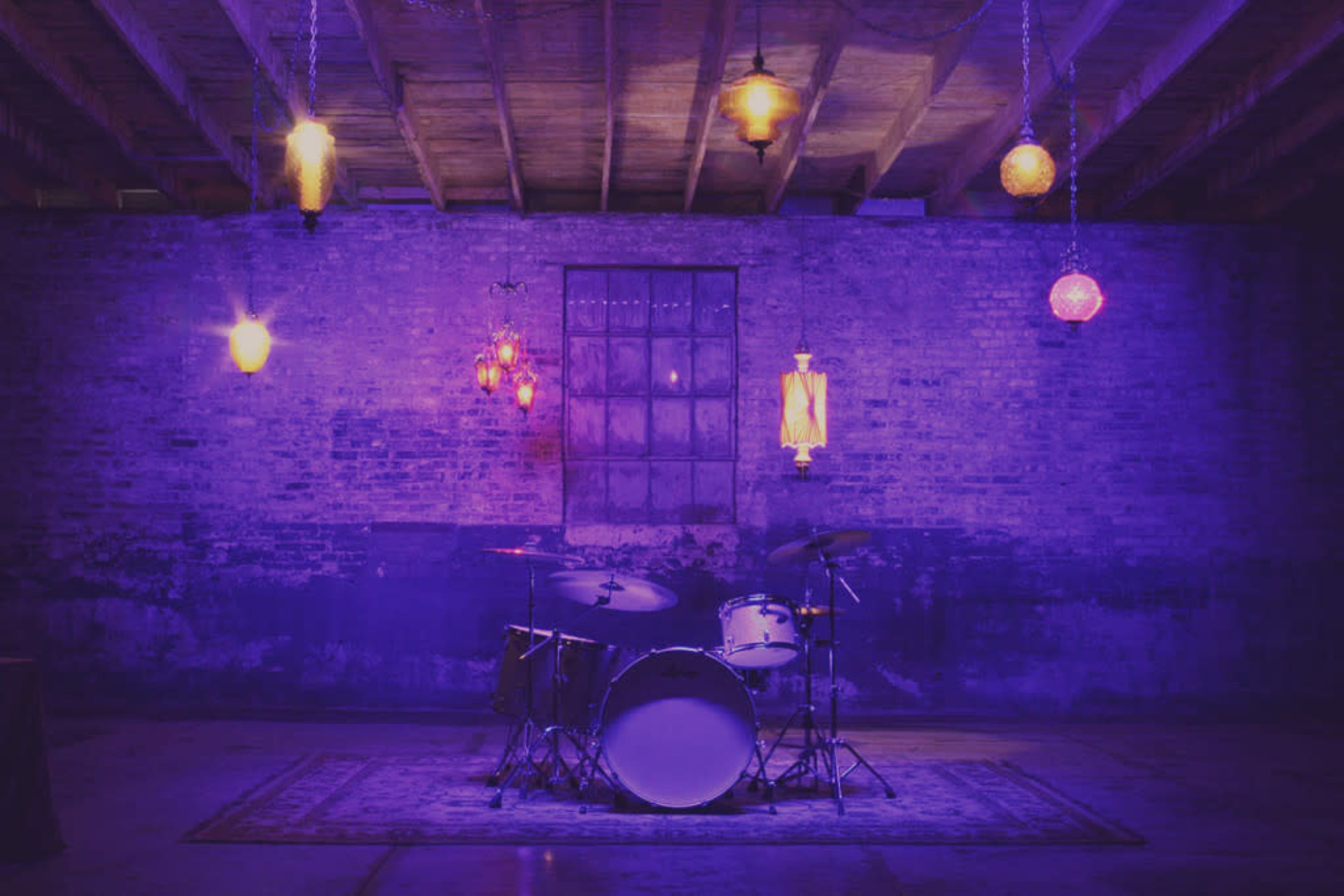 A drum set is positioned in front of a textured brick wall under various hanging lights creating a dimly lit atmosphere.