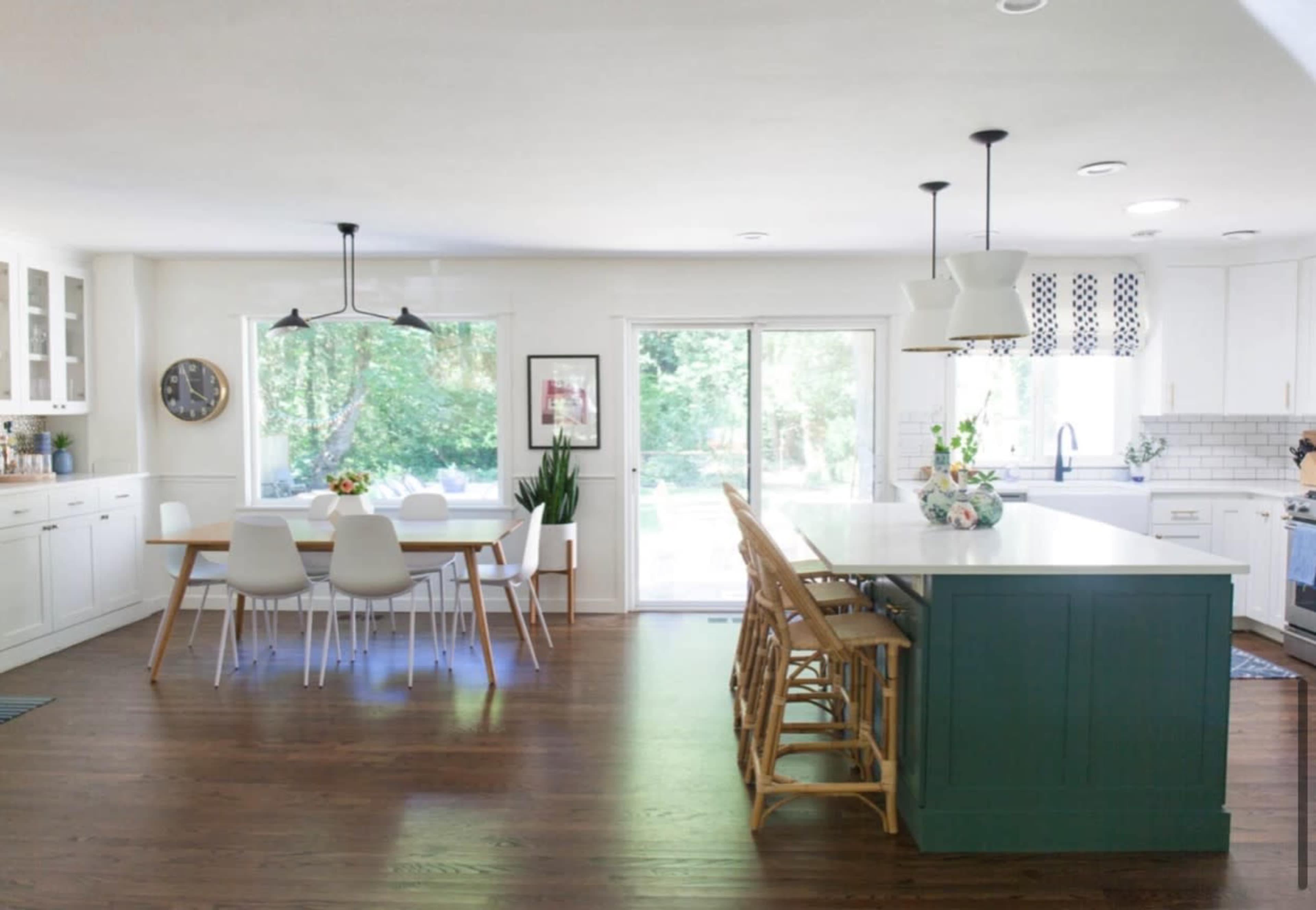 The kitchen features a large island with bar seating, a dining table, and a sliding glass door leading to a backyard, all surrounded by white cabinetry and natural wood floors.