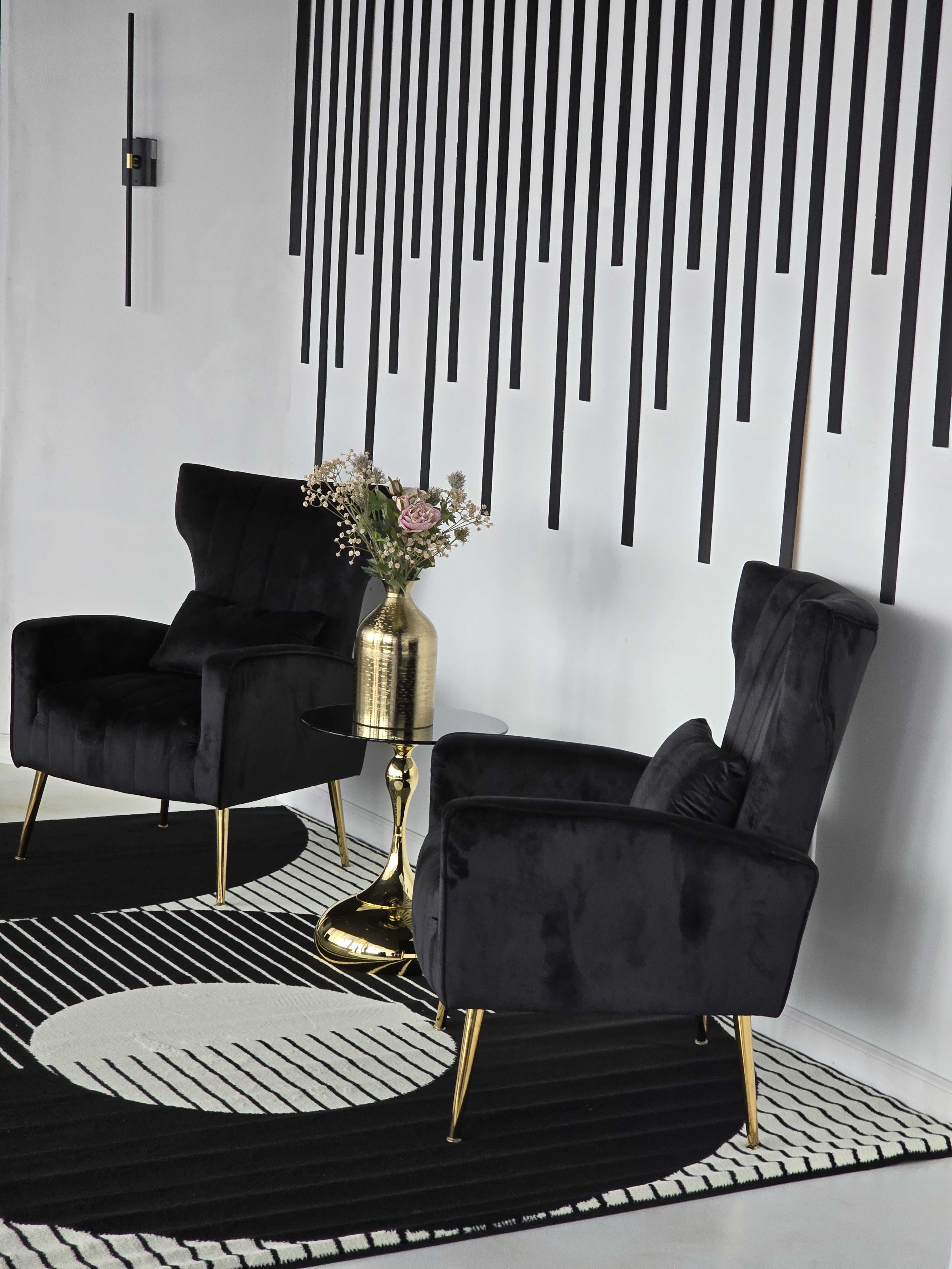 The image features two black velvet armchairs with gold legs flanking a small round table topped with a vase of flowers, set against a wall with vertical black stripes.