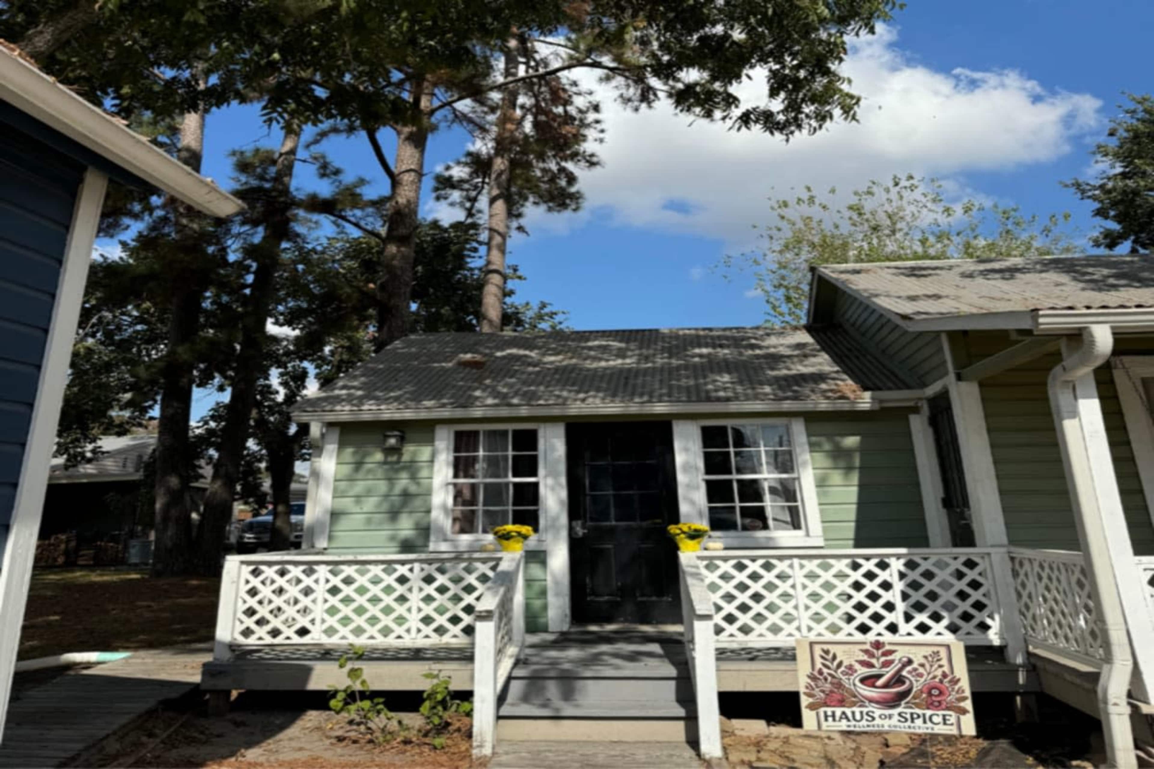 A small green house with a black door and white lattice railing is situated among tall trees, featuring a sign that reads "Haus of Spice."