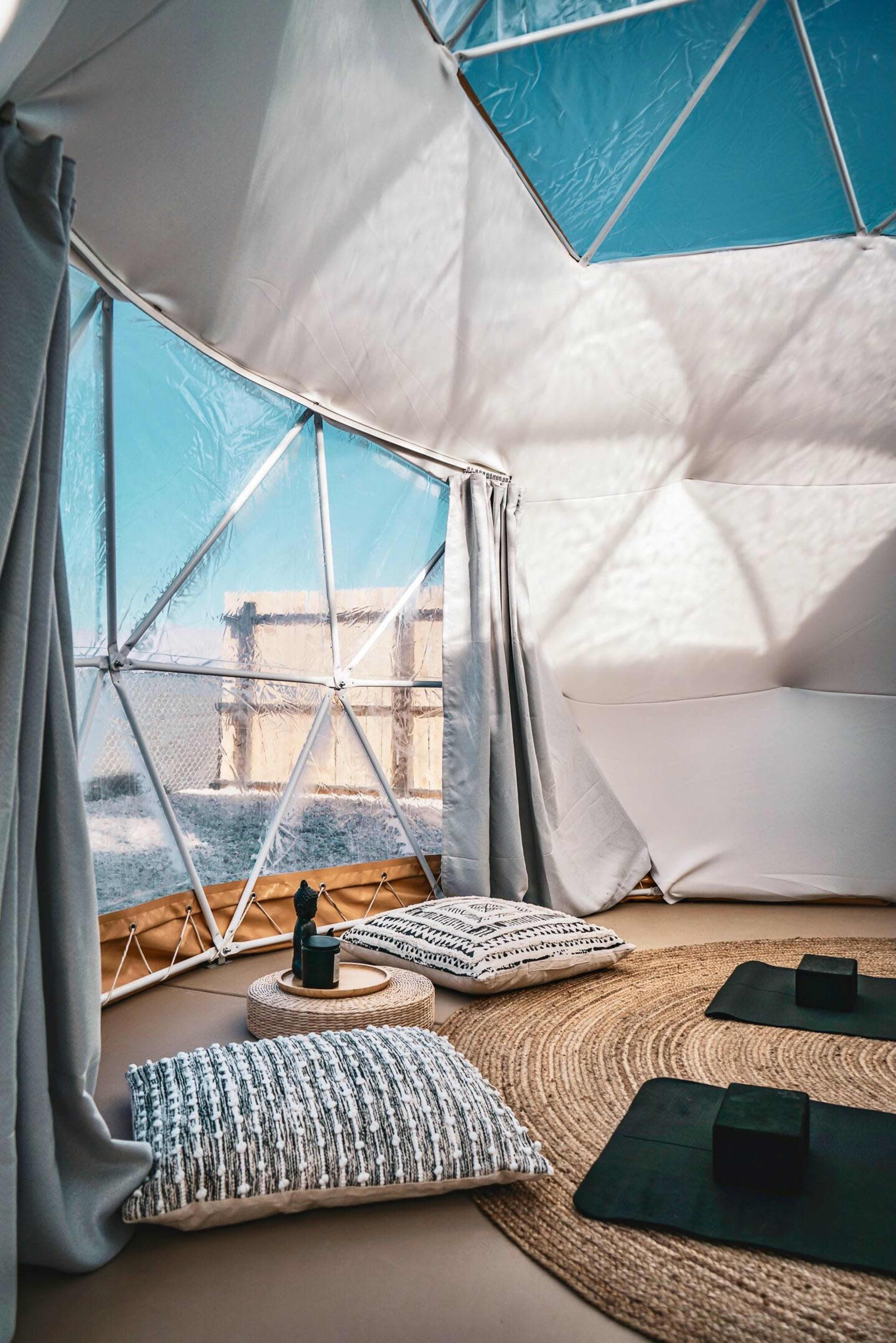The interior of a geodesic dome features floor cushions and a round rug, with large windows showcasing a clear sky outside.