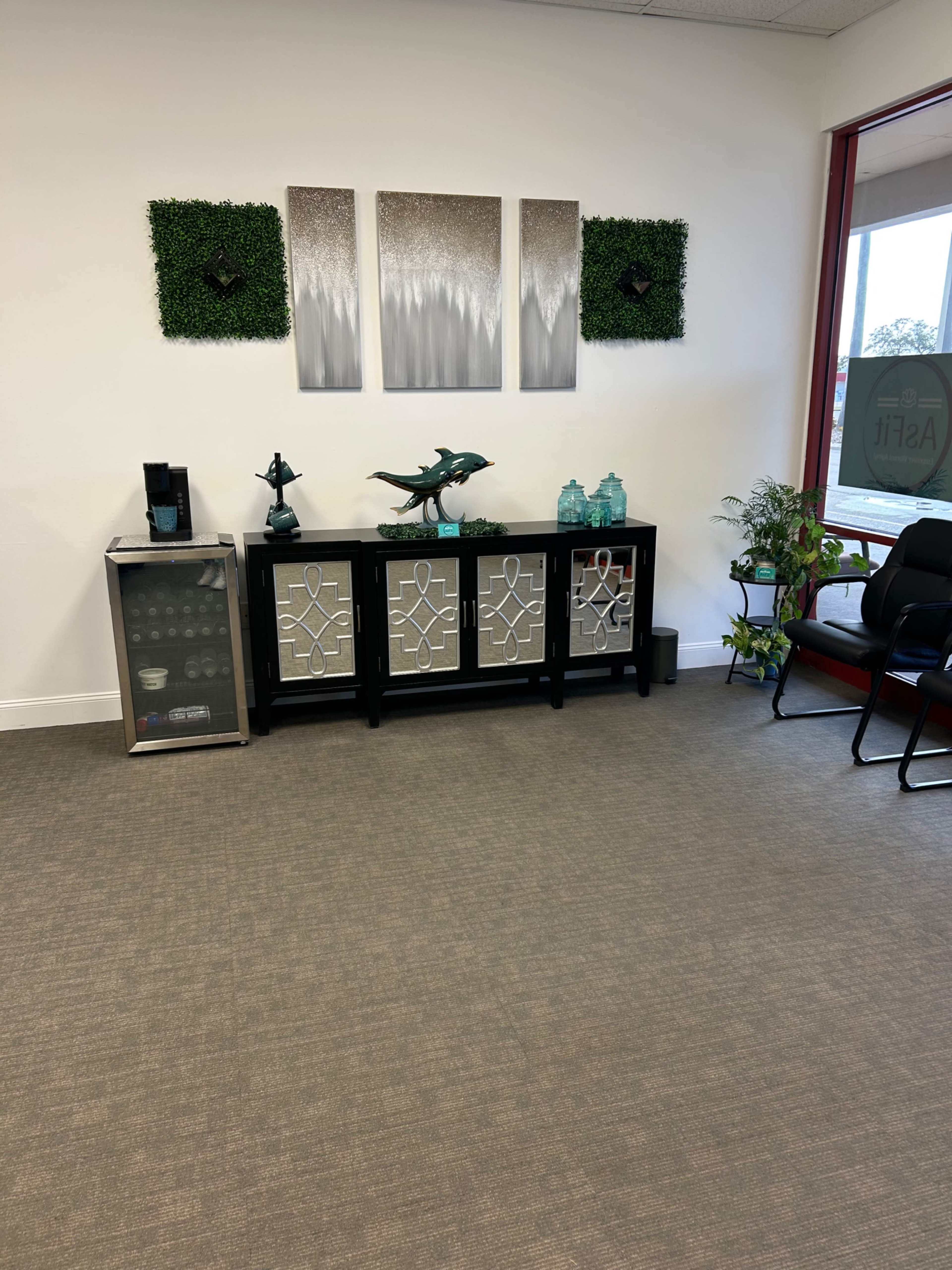 The image shows a modern office waiting area with a decorative black cabinet, wall art, plants, and a beverage station.