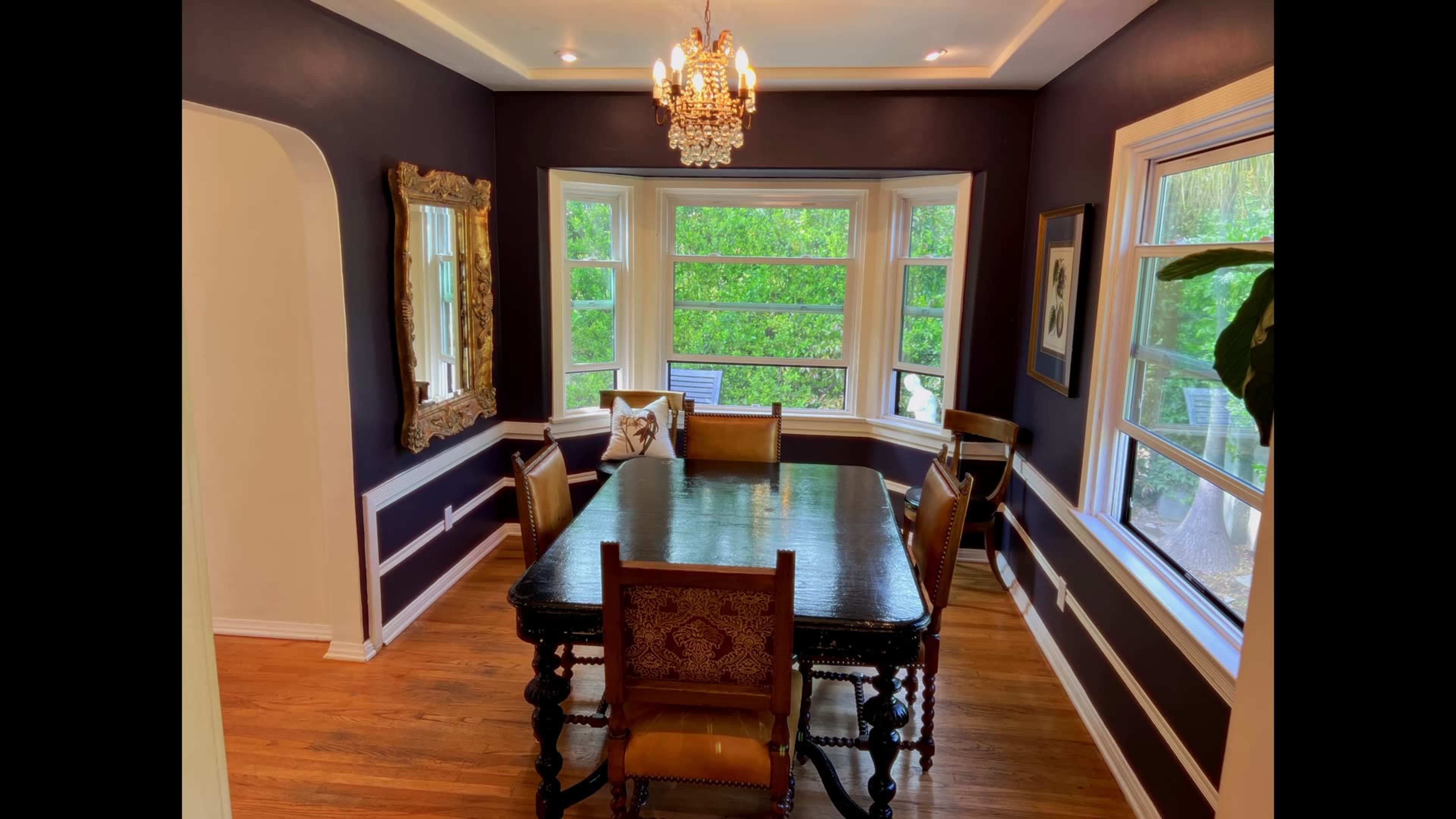 The image shows a dining room featuring a dark wood table surrounded by six chairs, a large mirror on the wall, and a chandelier hanging from the ceiling near a bay window with views of greenery outside.