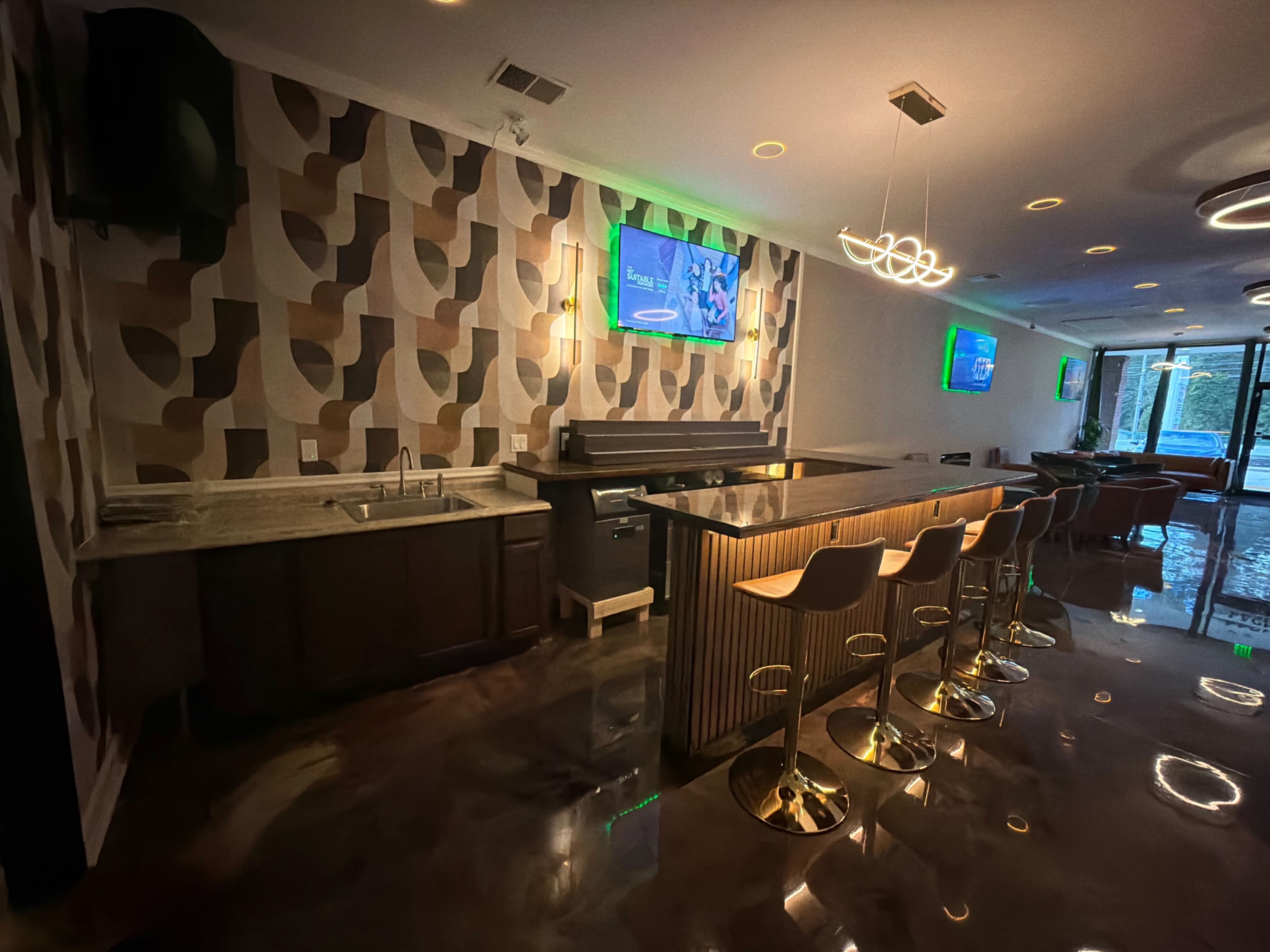 The image shows a modern bar area featuring a sleek countertop, high stools, and patterned wallpaper with illuminated screens on the walls.
