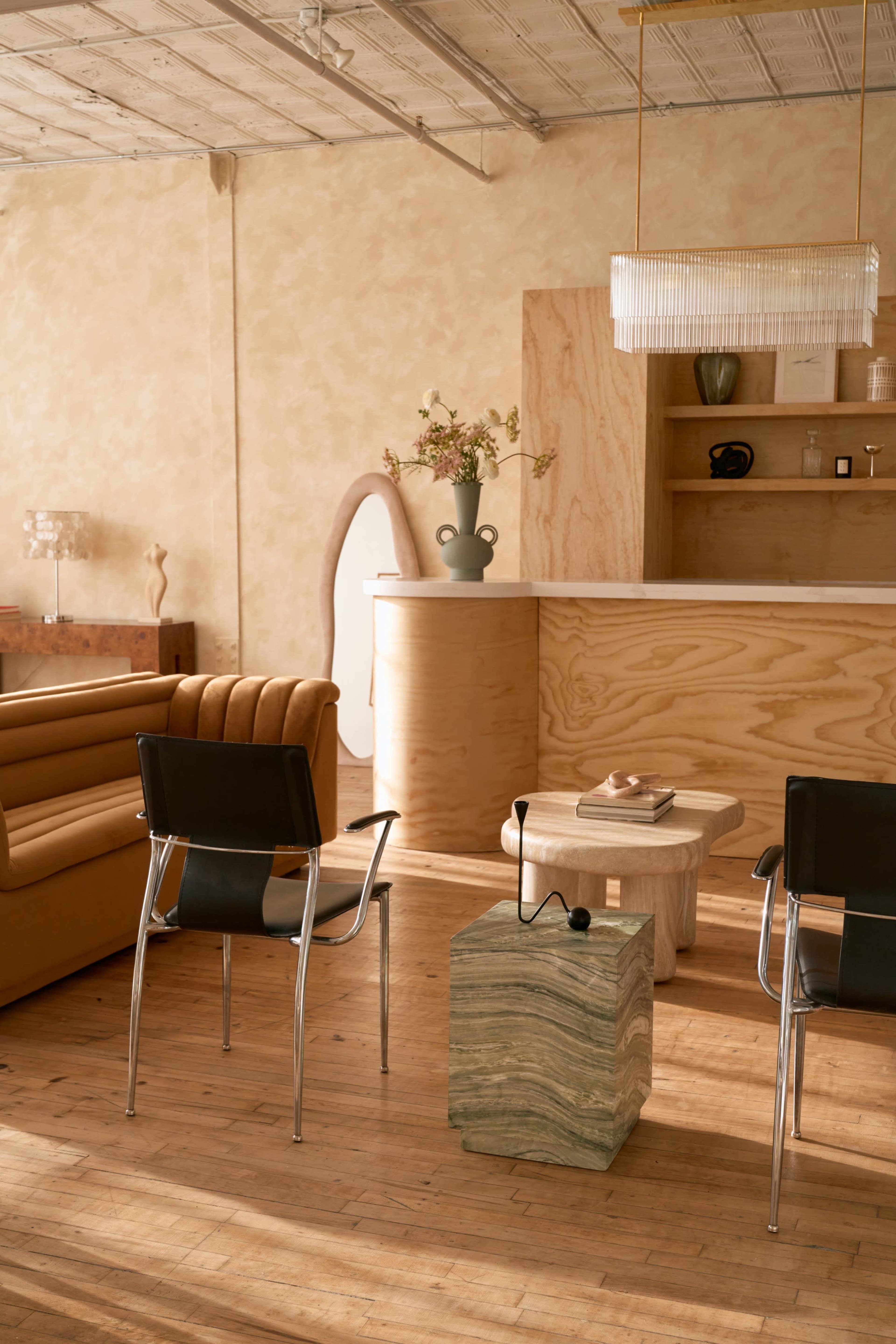 The image shows a modern interior with a wooden sofa, two black chairs, and a sculptural stone coffee table in a brightly lit space.
