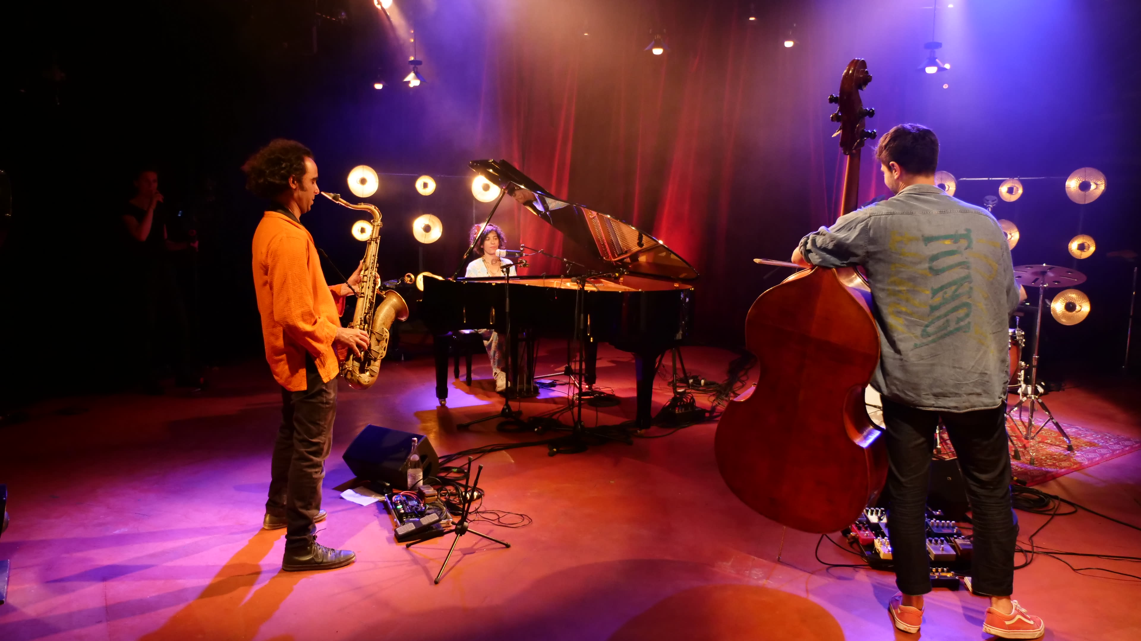 A trio of musicians performs on stage, with a saxophonist, a pianist, and a bassist, under vibrant stage lighting.