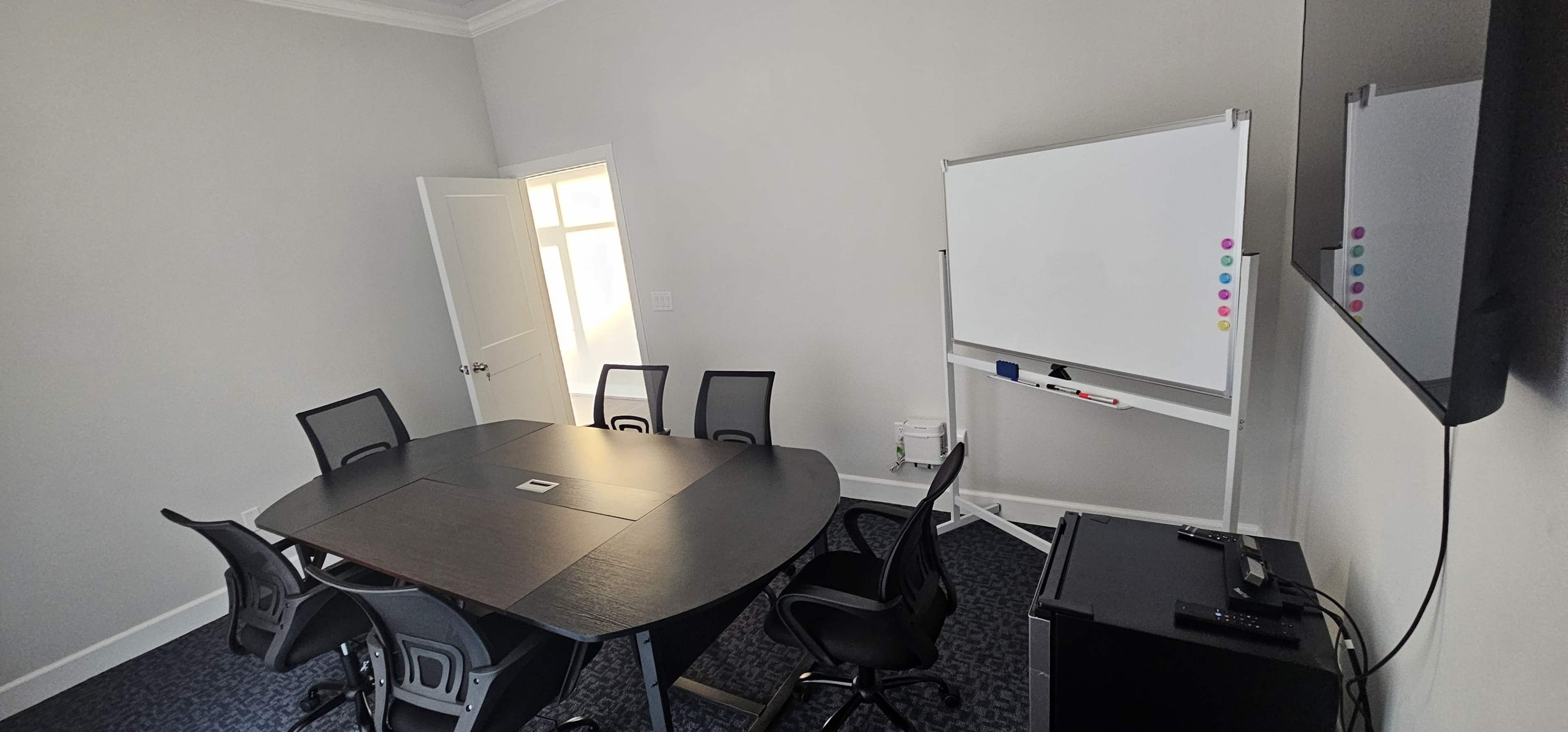 A conference room is arranged with a large table, six black chairs, a whiteboard on a stand, and a wall-mounted screen.
