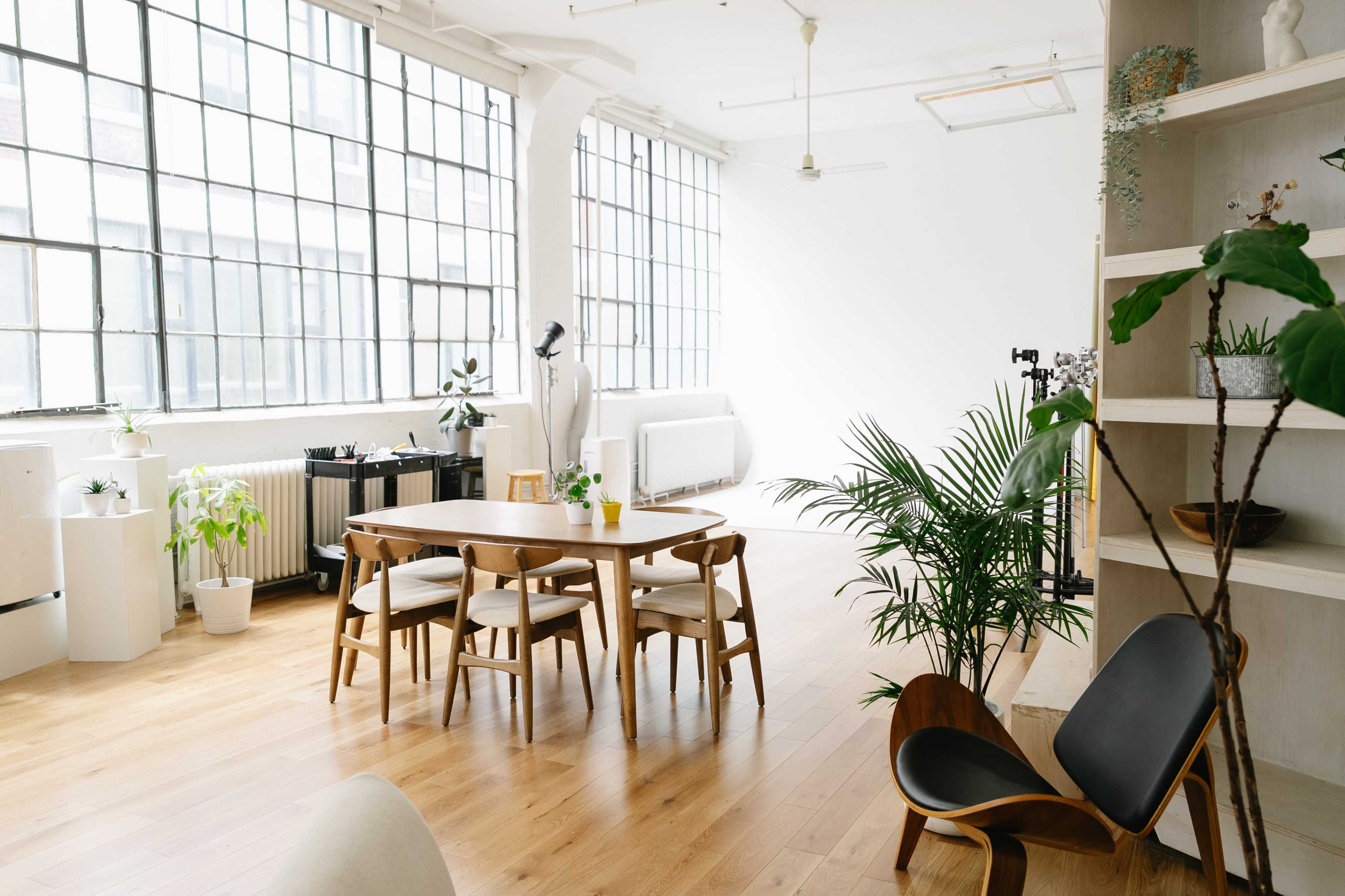 The image shows a spacious, modern studio with large windows, wooden flooring, and a dining table surrounded by chairs, along with plants and furniture.