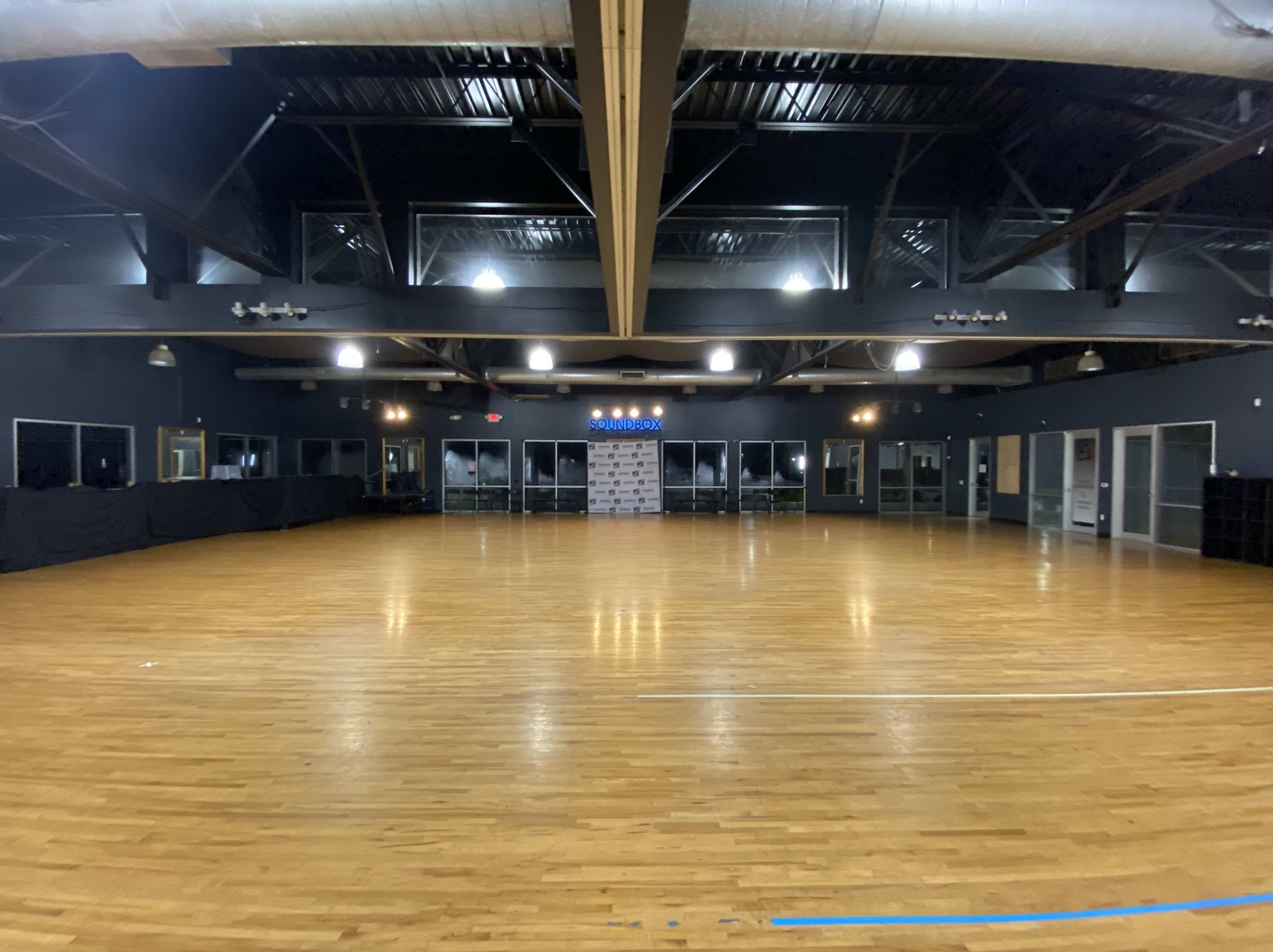 A large, empty hardwood-floored room with high ceilings and a wall of windows at one end.