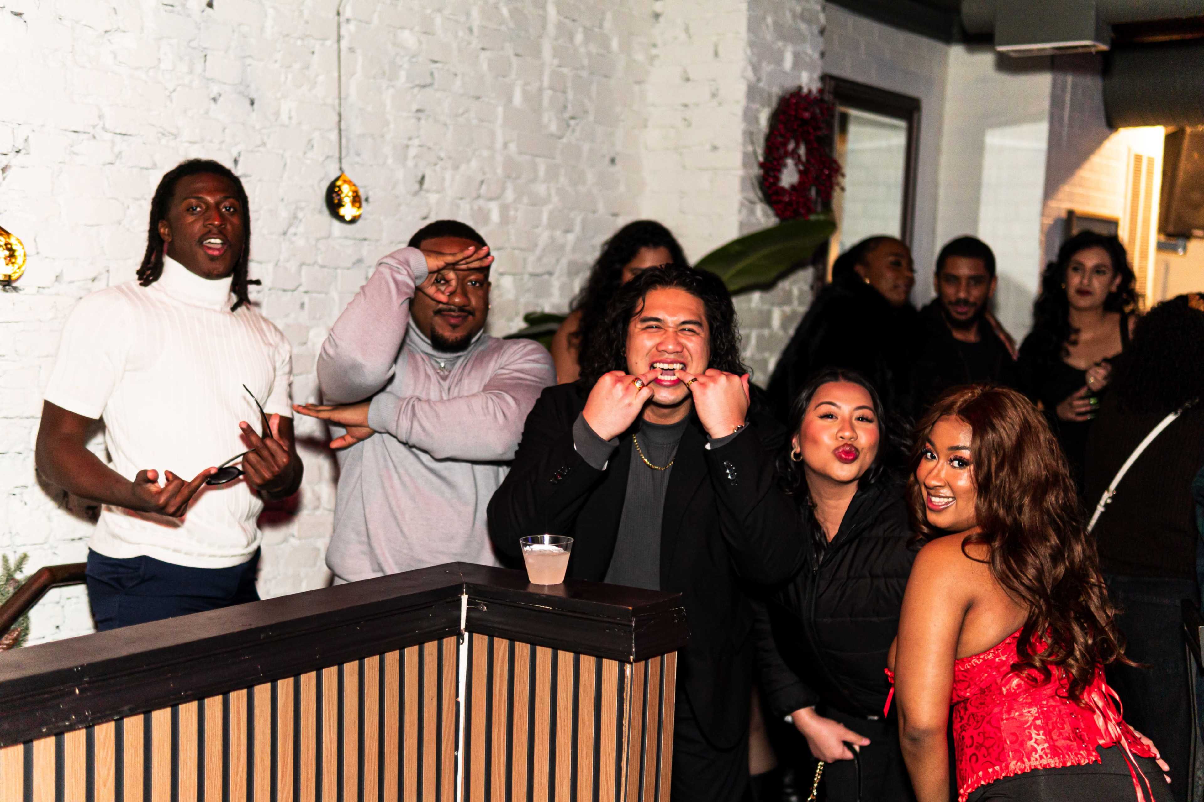 A group of people poses playfully in a social setting, with some making various gestures and facial expressions.