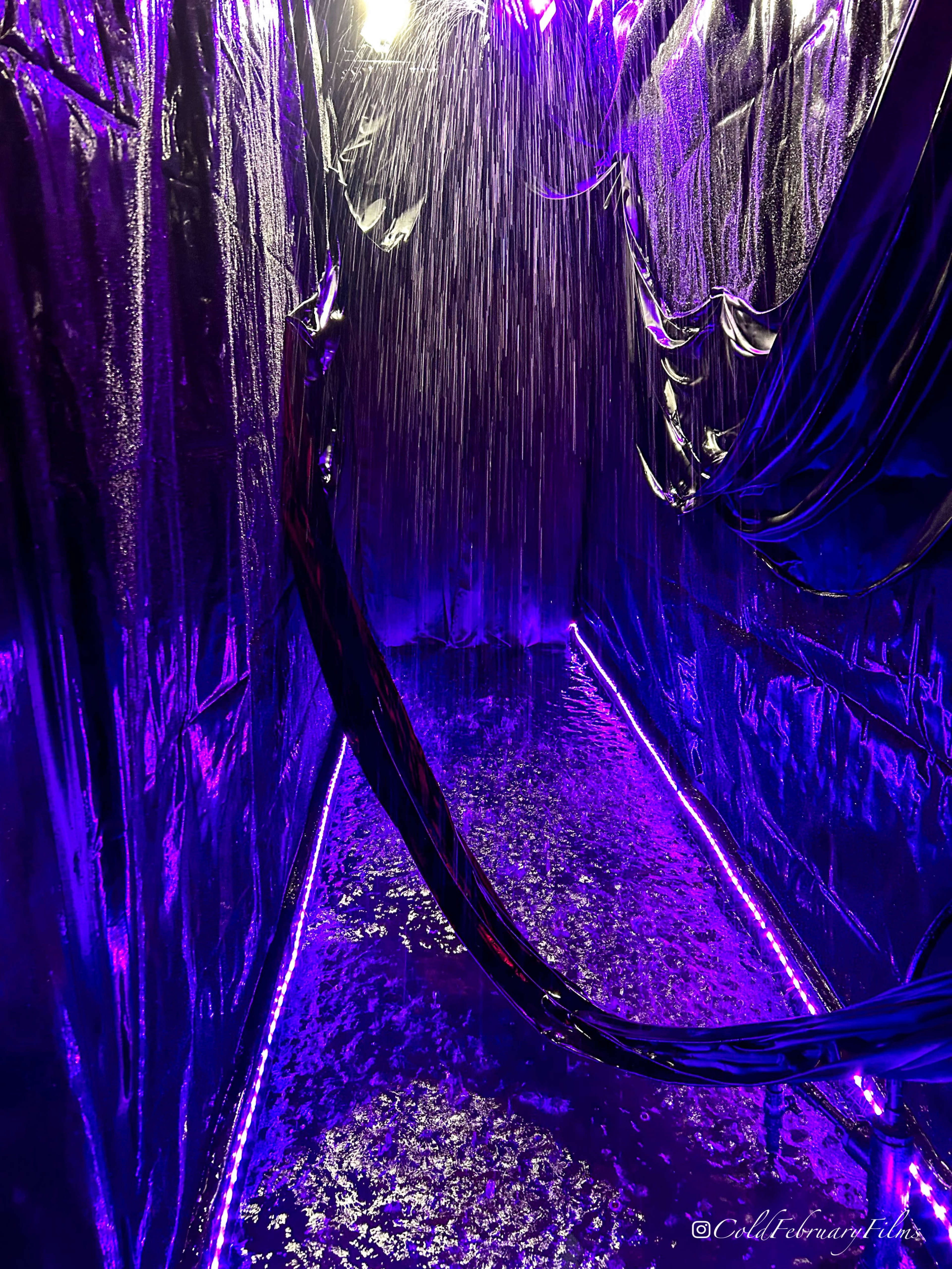 The image shows a narrow corridor with reflective surfaces and illuminated purple lights, featuring water cascading from above.