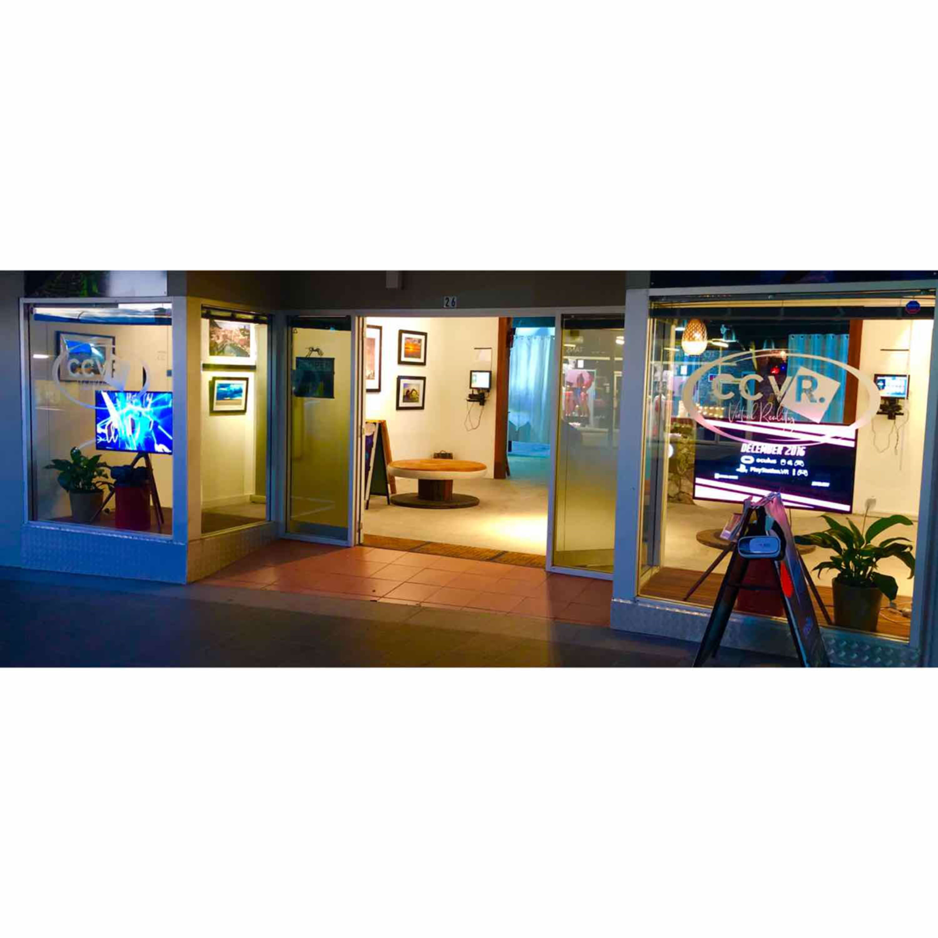 The exterior of a gallery with glass doors, displaying art and digital screens inside, framed by potted plants.