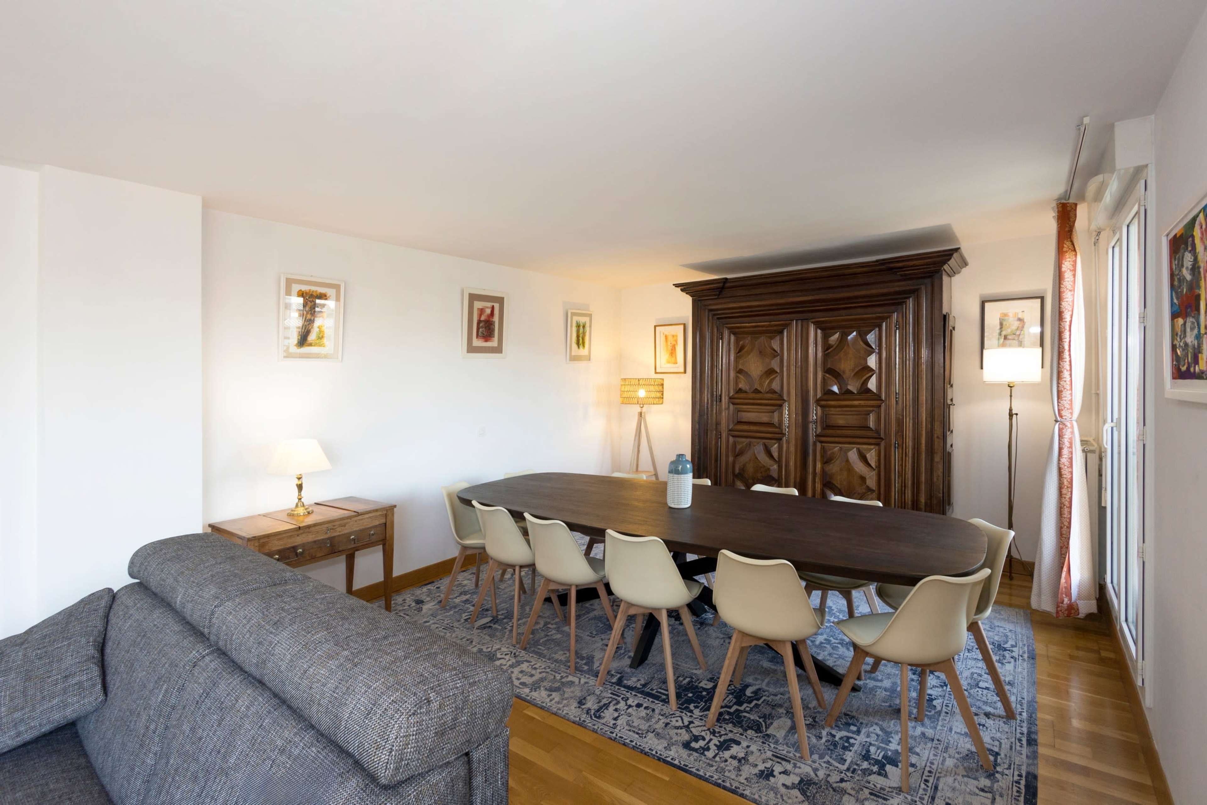 The image shows a dining room with a large wooden table surrounded by chairs, an ornate cabinet, and various wall art.